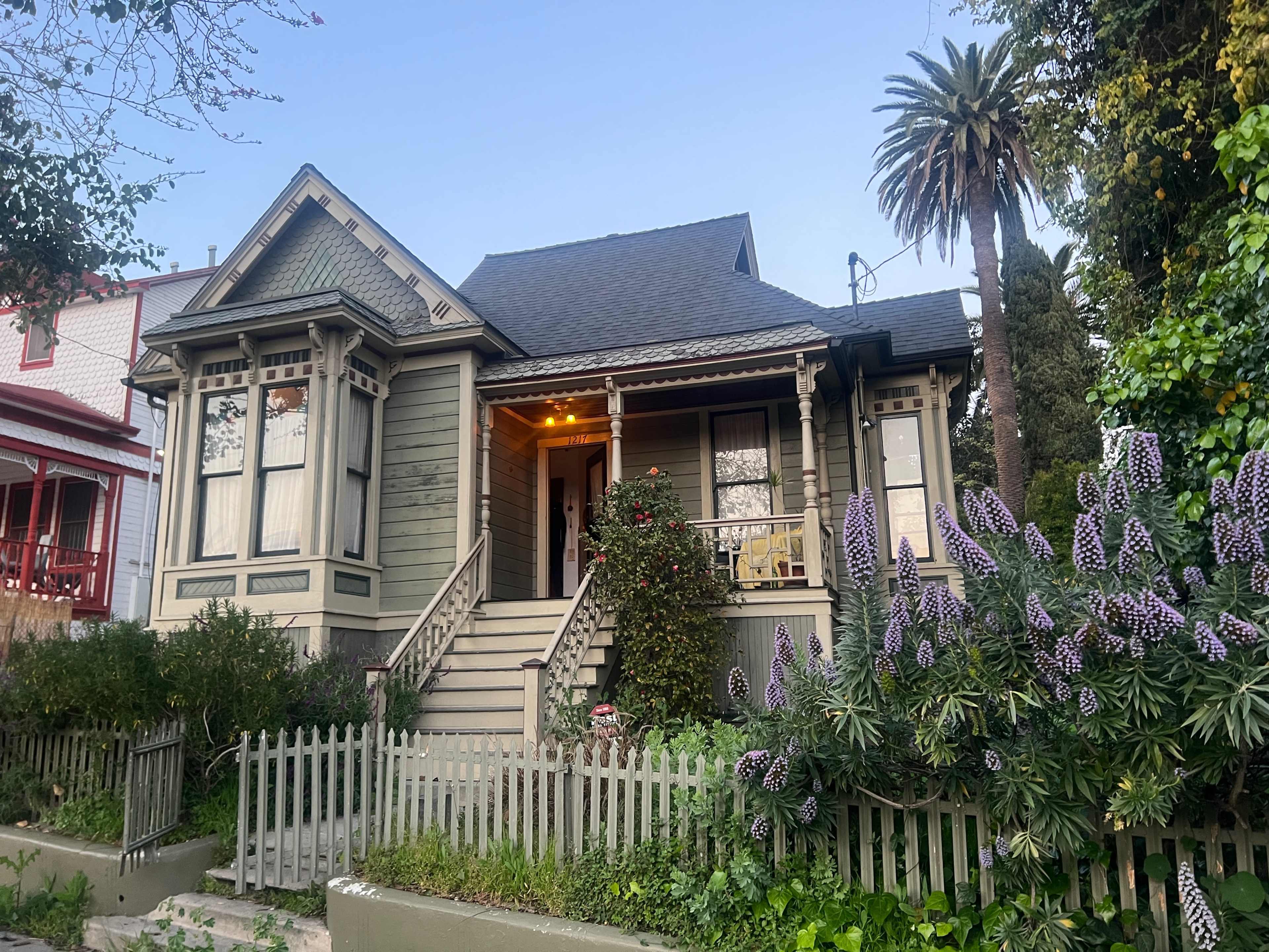 A two-story Victorian-style house with a front porch, surrounded by green foliage and blooming flowers, is located on a well-maintained property.