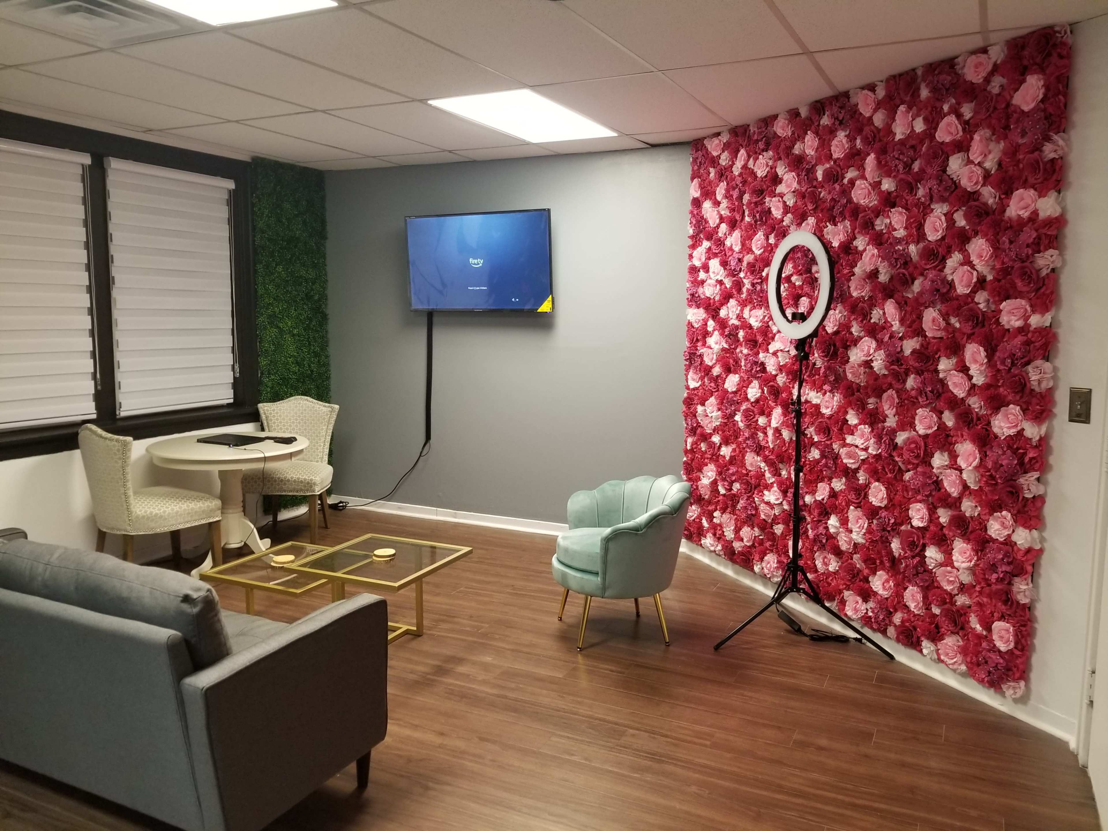 A modern lounge area with a gray sofa, light blue armchair, table, and a large wall covered in pink and white artificial flowers.