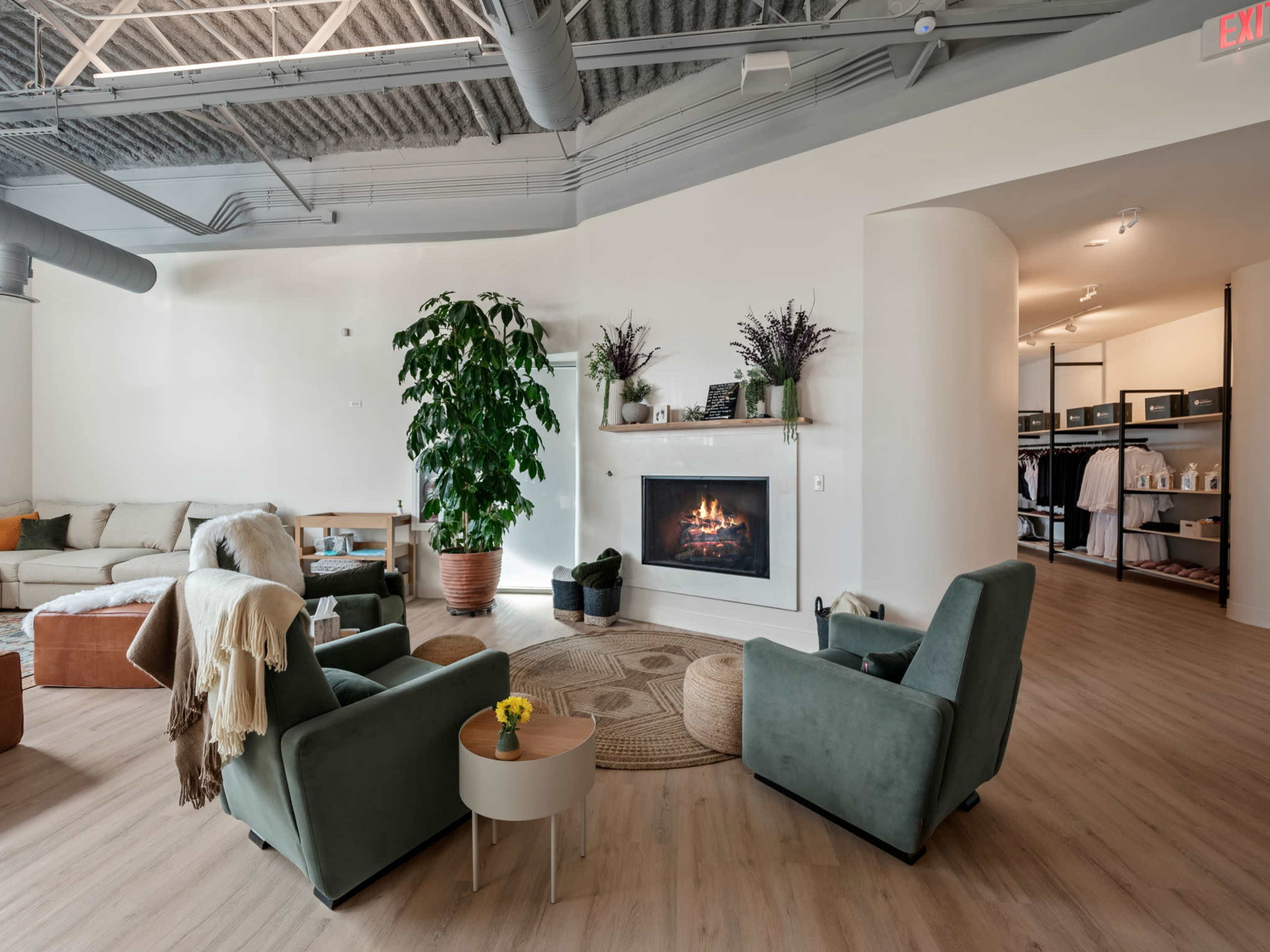 A modern living room features two green armchairs facing a white fireplace, with a large indoor plant and a cozy seating area in the background.