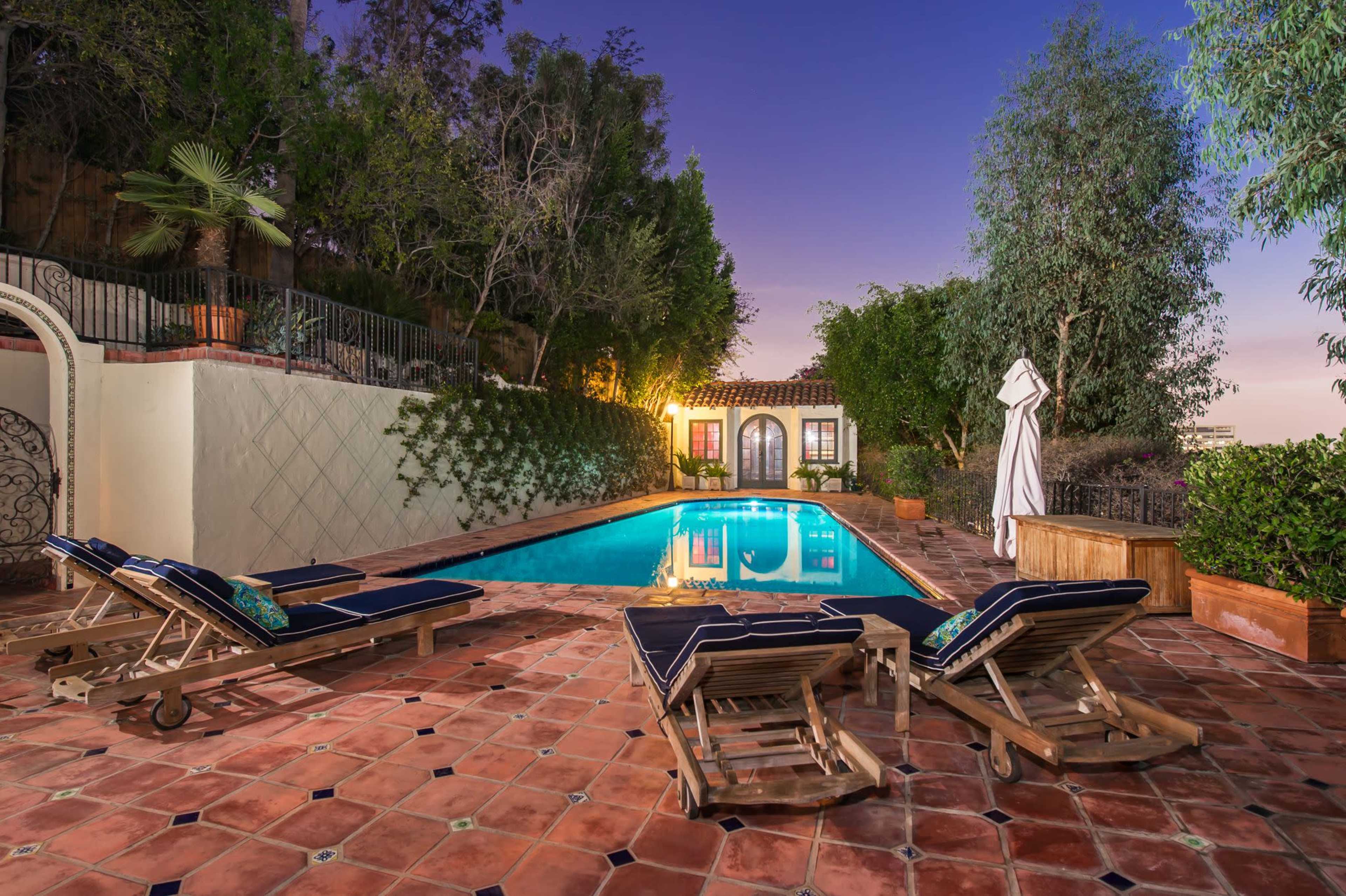 The image shows a tranquil outdoor pool area with lounge chairs, surrounded by greenery and a gated wall, illuminated by evening lights.
