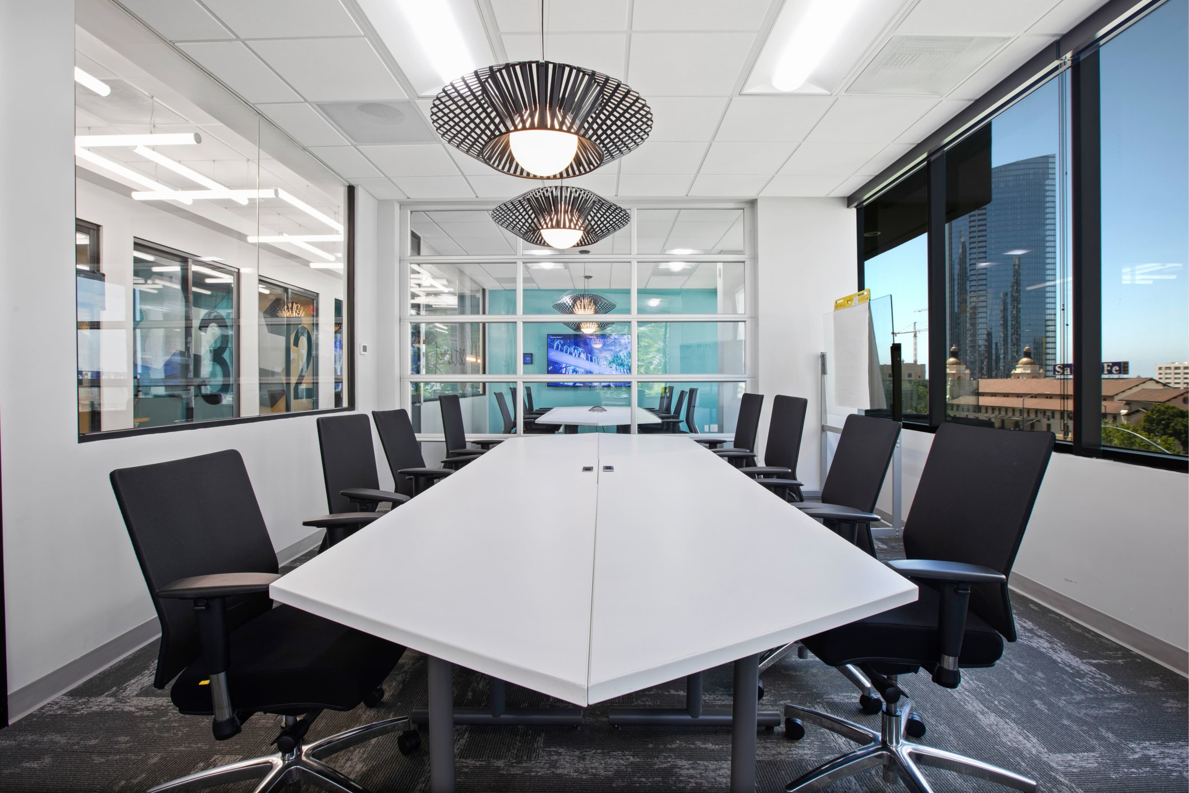 A modern conference room features a long, white table surrounded by black chairs, with large windows providing a view of a cityscape.