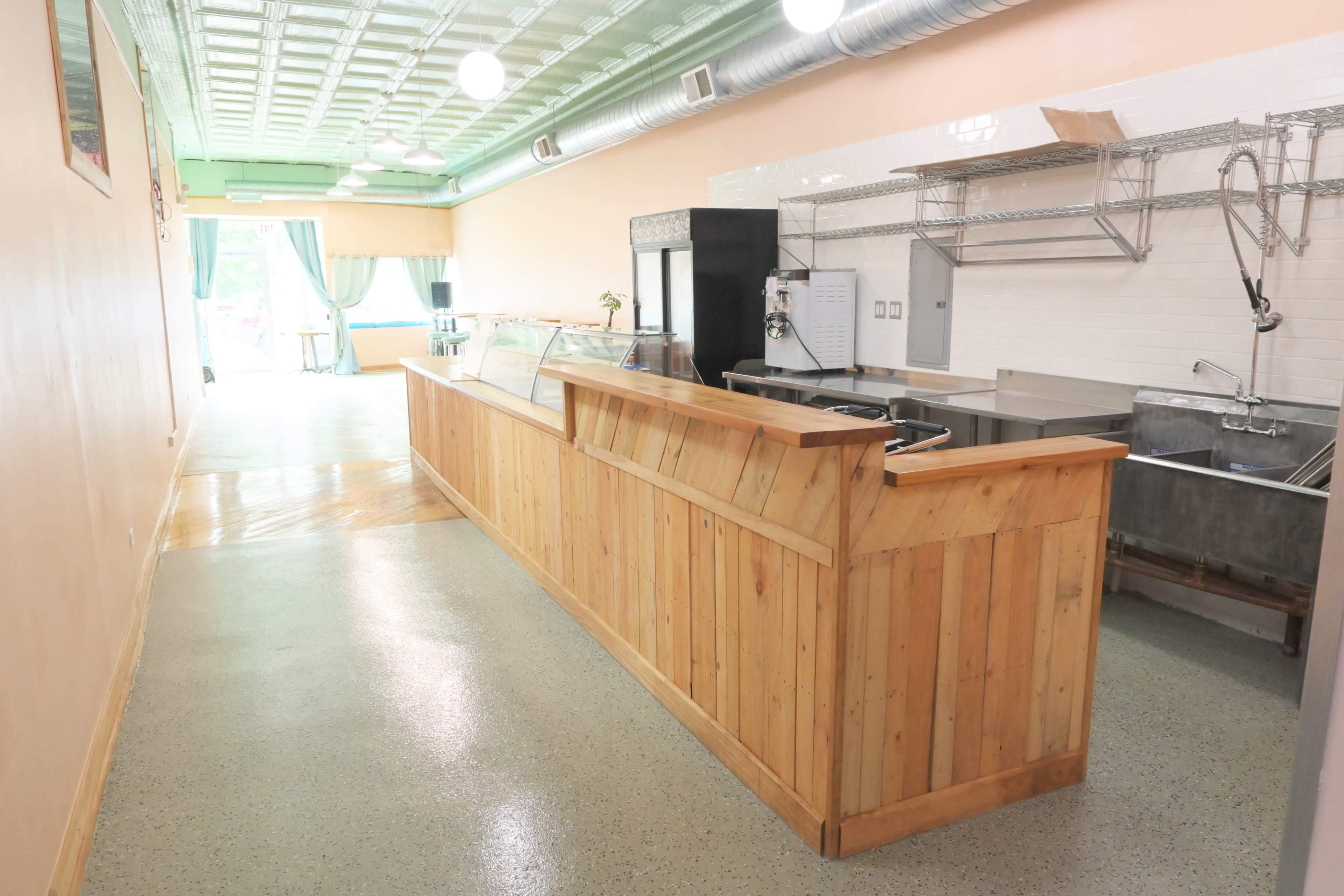A spacious, empty café interior featuring a wooden counter with glass display cases, stainless steel sinks, and a tiled wall.