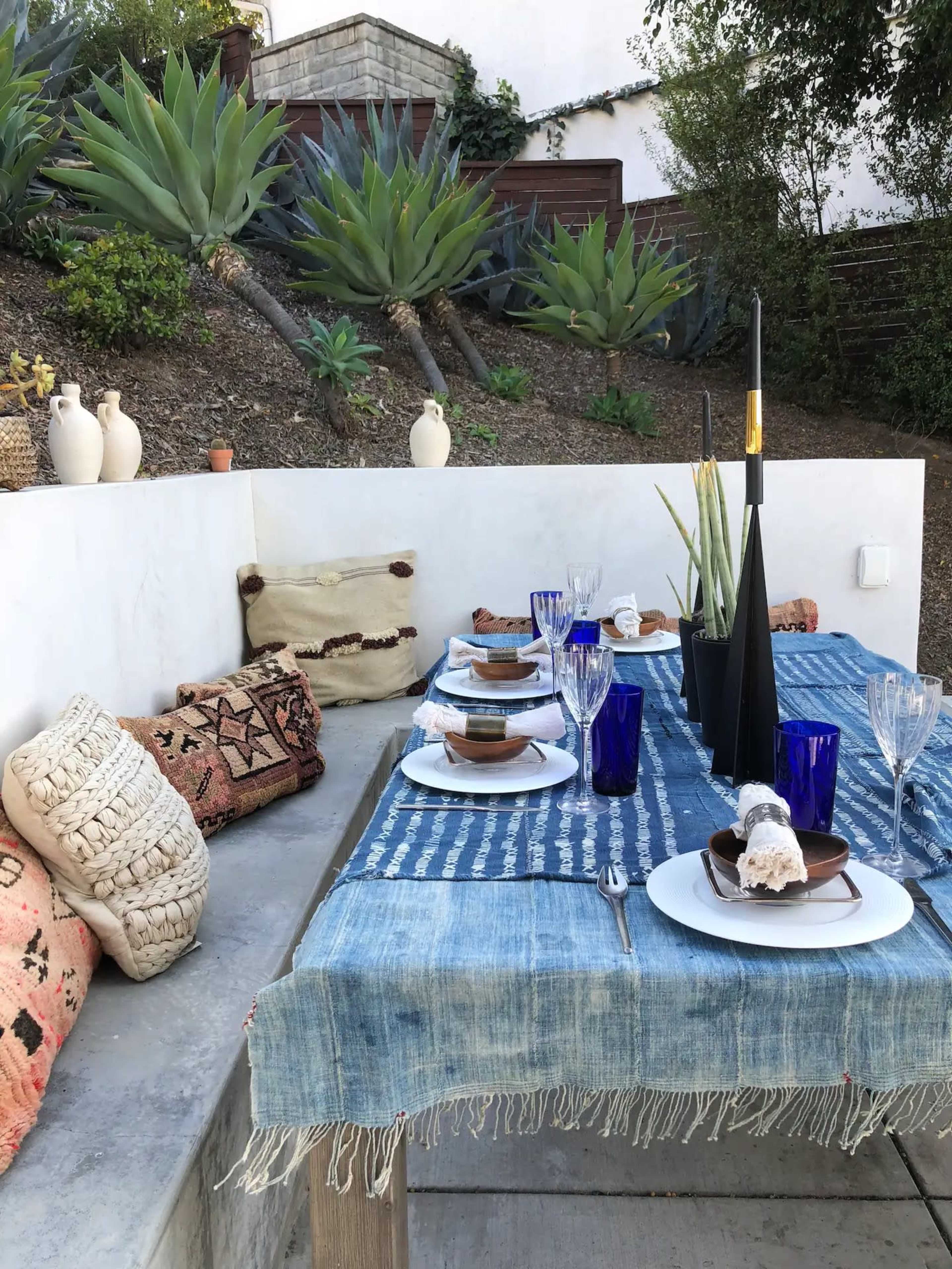 A rustic dining table is set with blue glassware and plates, surrounded by decorative cushions, against a backdrop of greenery and terraced landscaping.