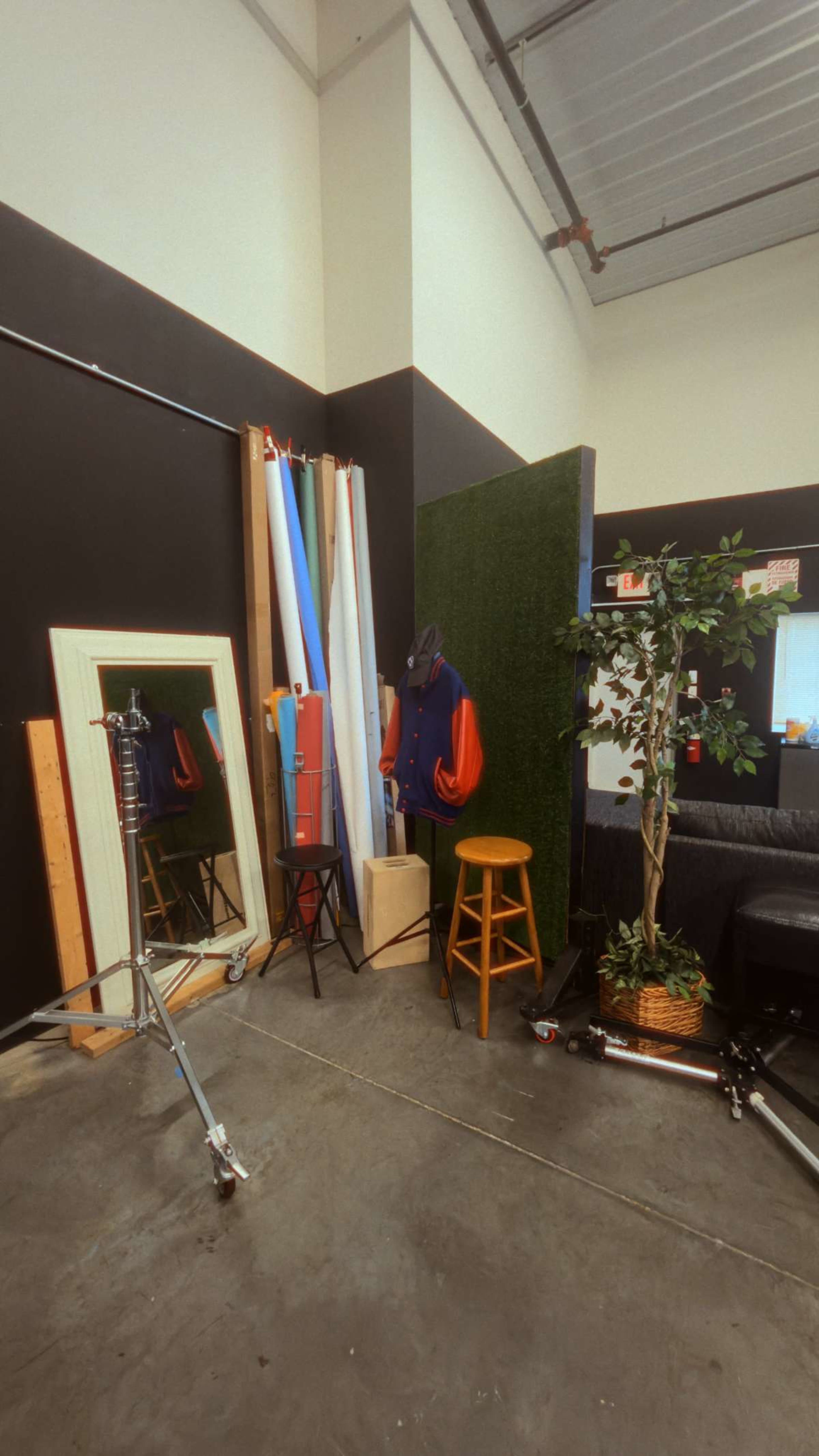 The image shows a corner of a studio featuring a mirror, backdrop materials, a garment on a mannequin, a stool, a potted plant, and a black couch.
