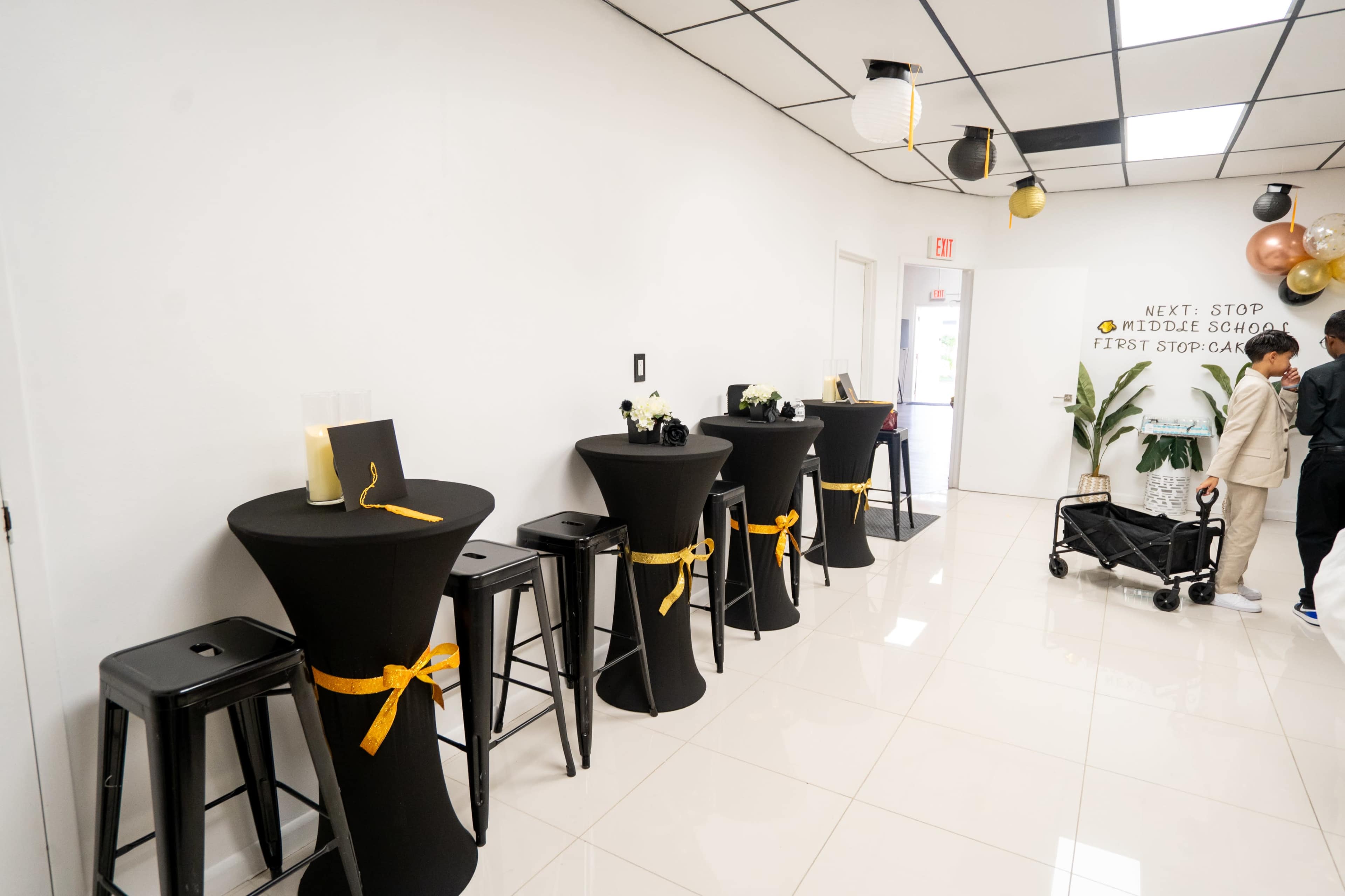 The image shows a white-walled room with several black cocktail tables, each adorned with a yellow ribbon, and a person pushing a stroller near the entrance.