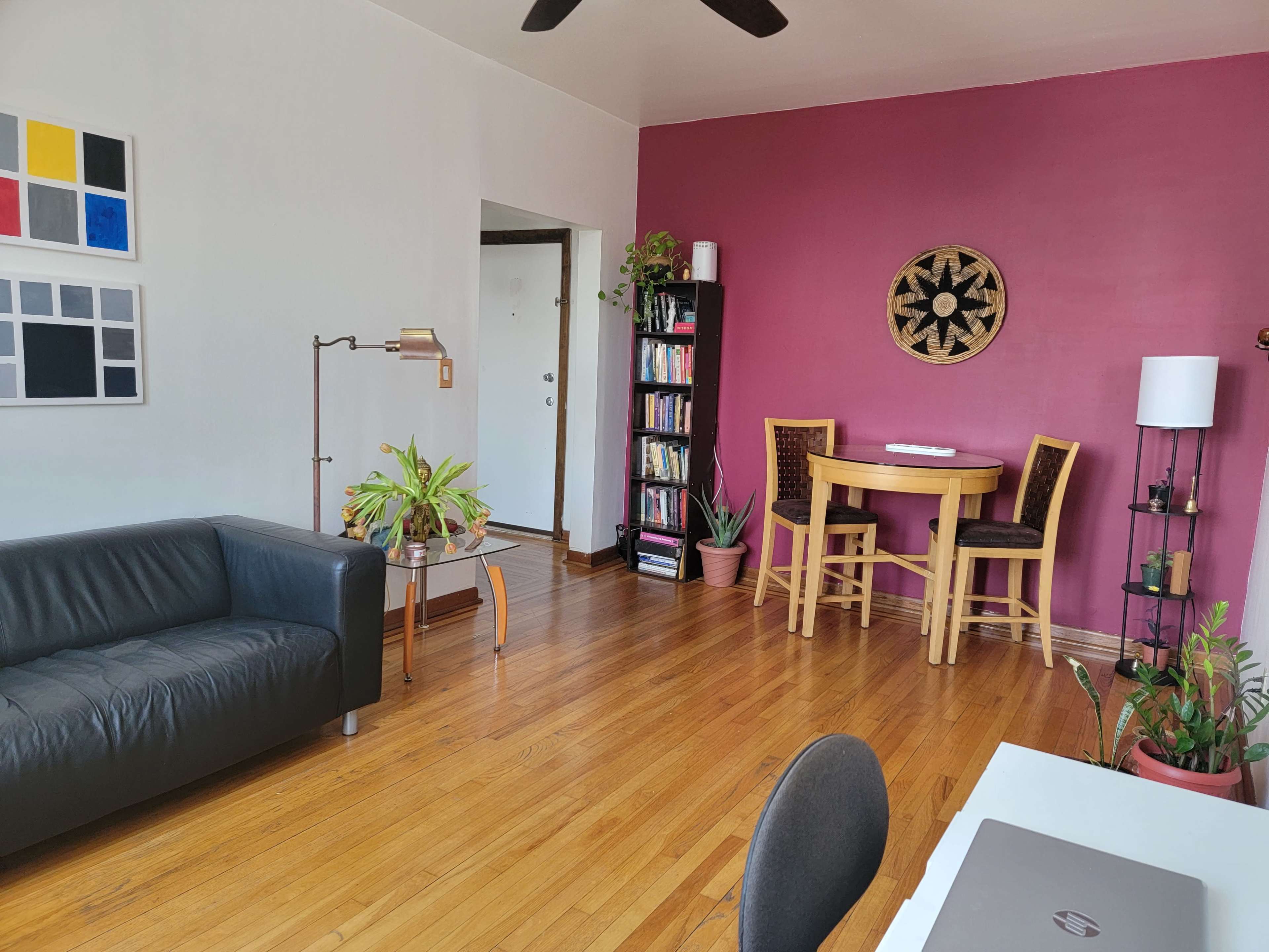 A living space with a black leather sofa, a round dining table with chairs, bookshelves filled with books, and several potted plants against a pink wall.