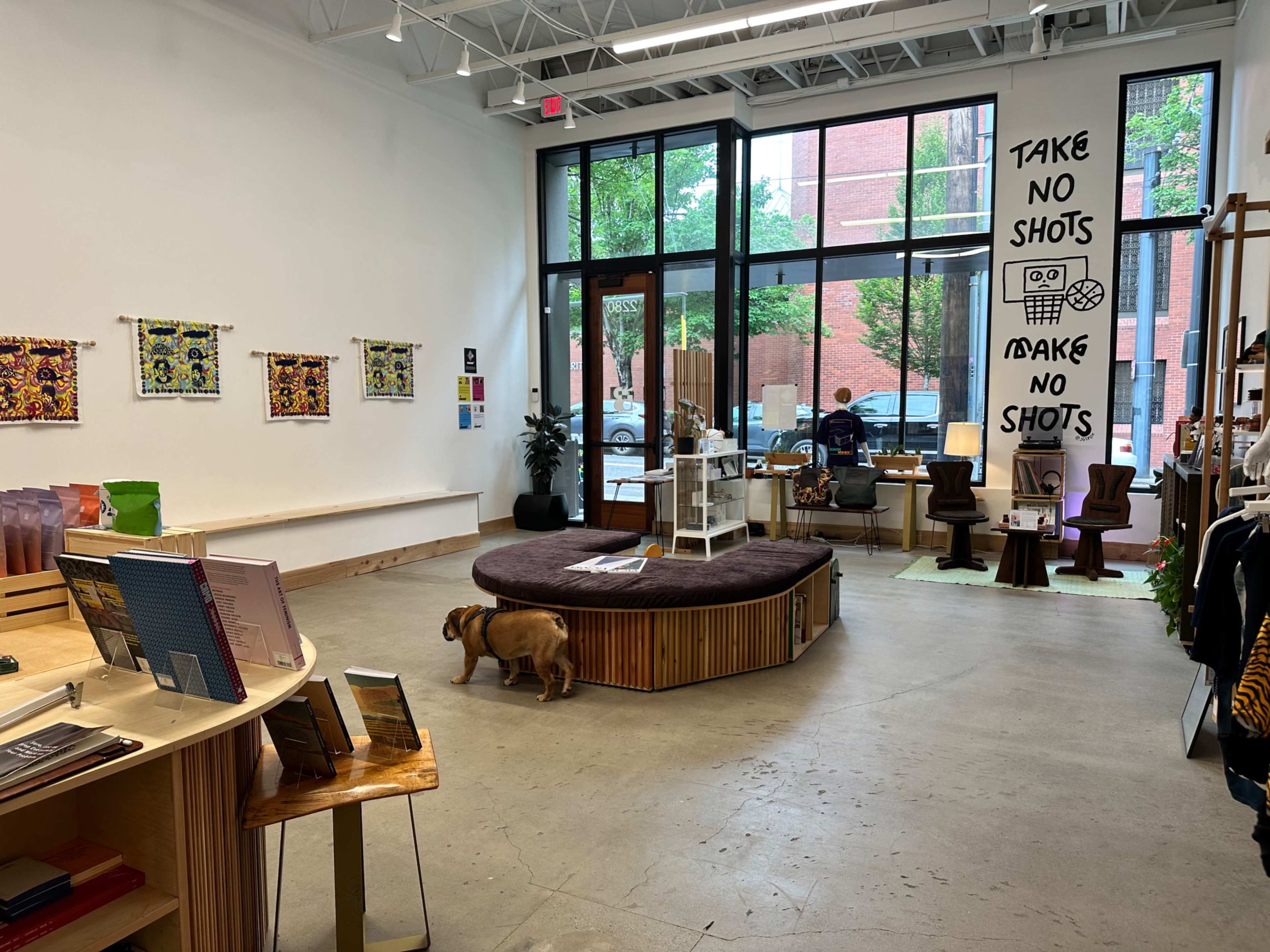 A spacious retail store features a circular seating area and displays of books and art alongside a large window, while a dog walks along the floor.