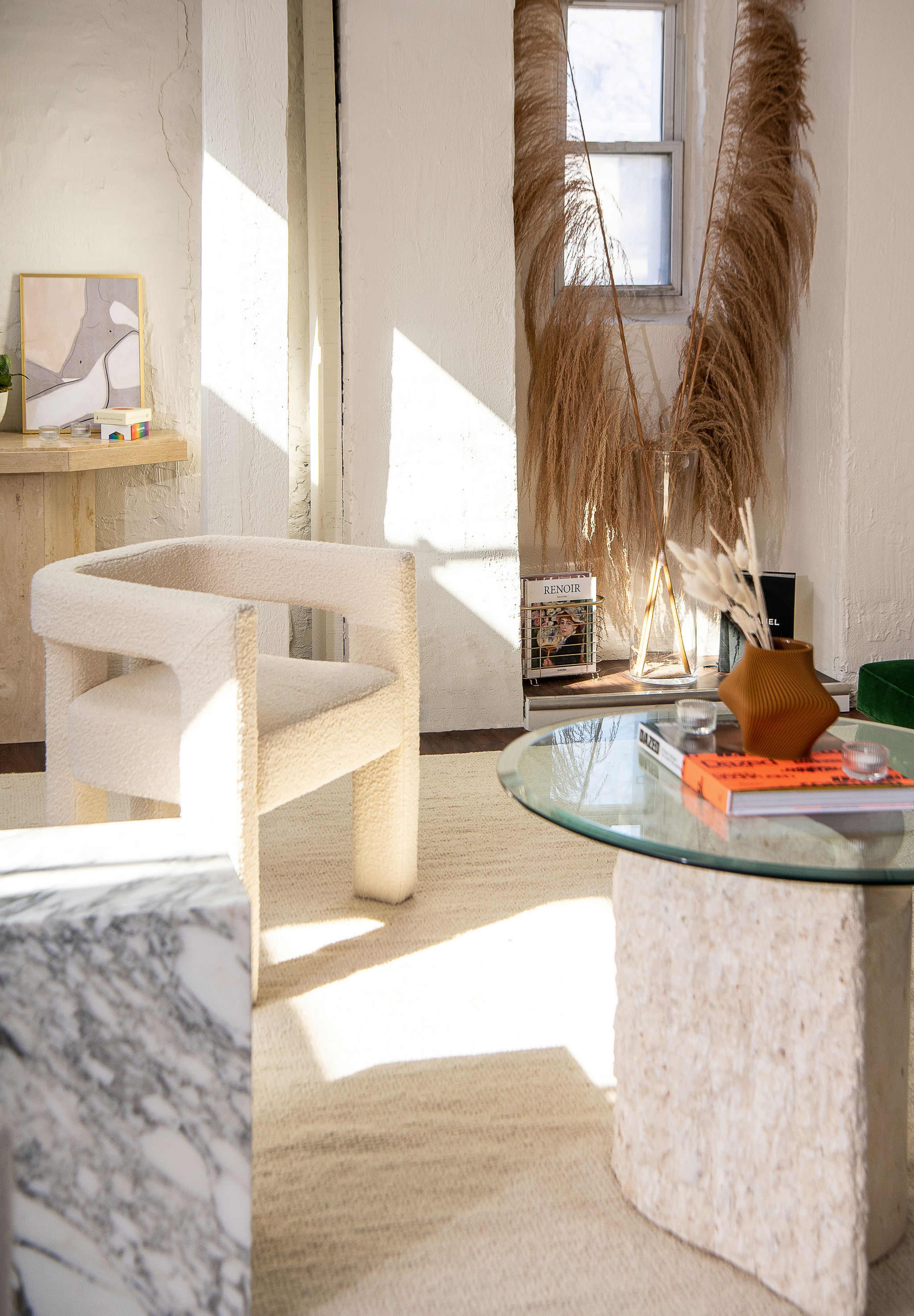 A modern interior space with a textural cream-colored chair, a glass-top table on a textured base, and decorative elements like pampas grass in a corner.