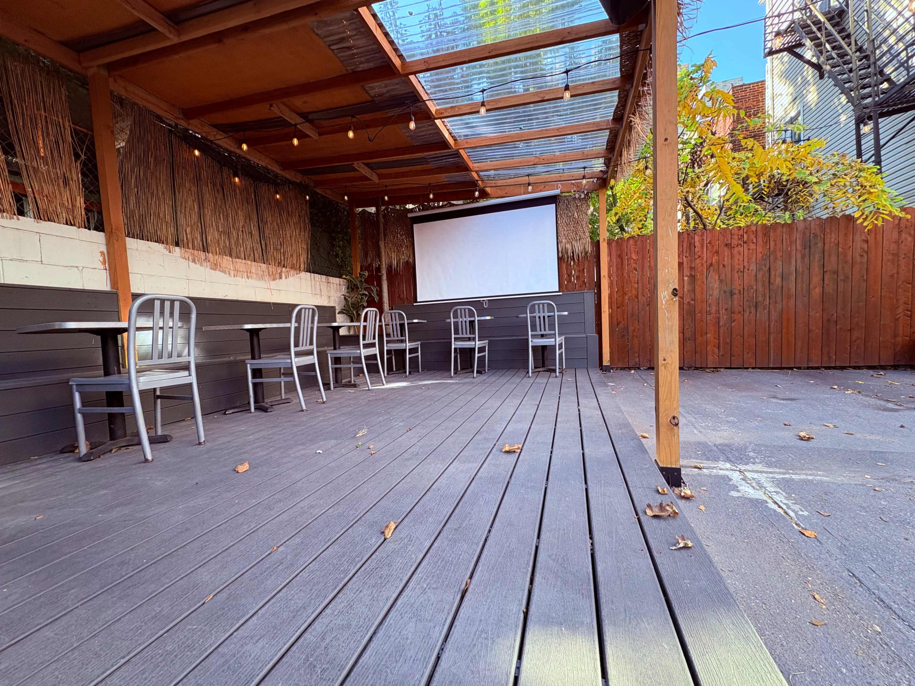 The image shows an empty outdoor seating area of a restaurant with wooden flooring, metal chairs arranged around tables, and a large screen against a wooden fence.