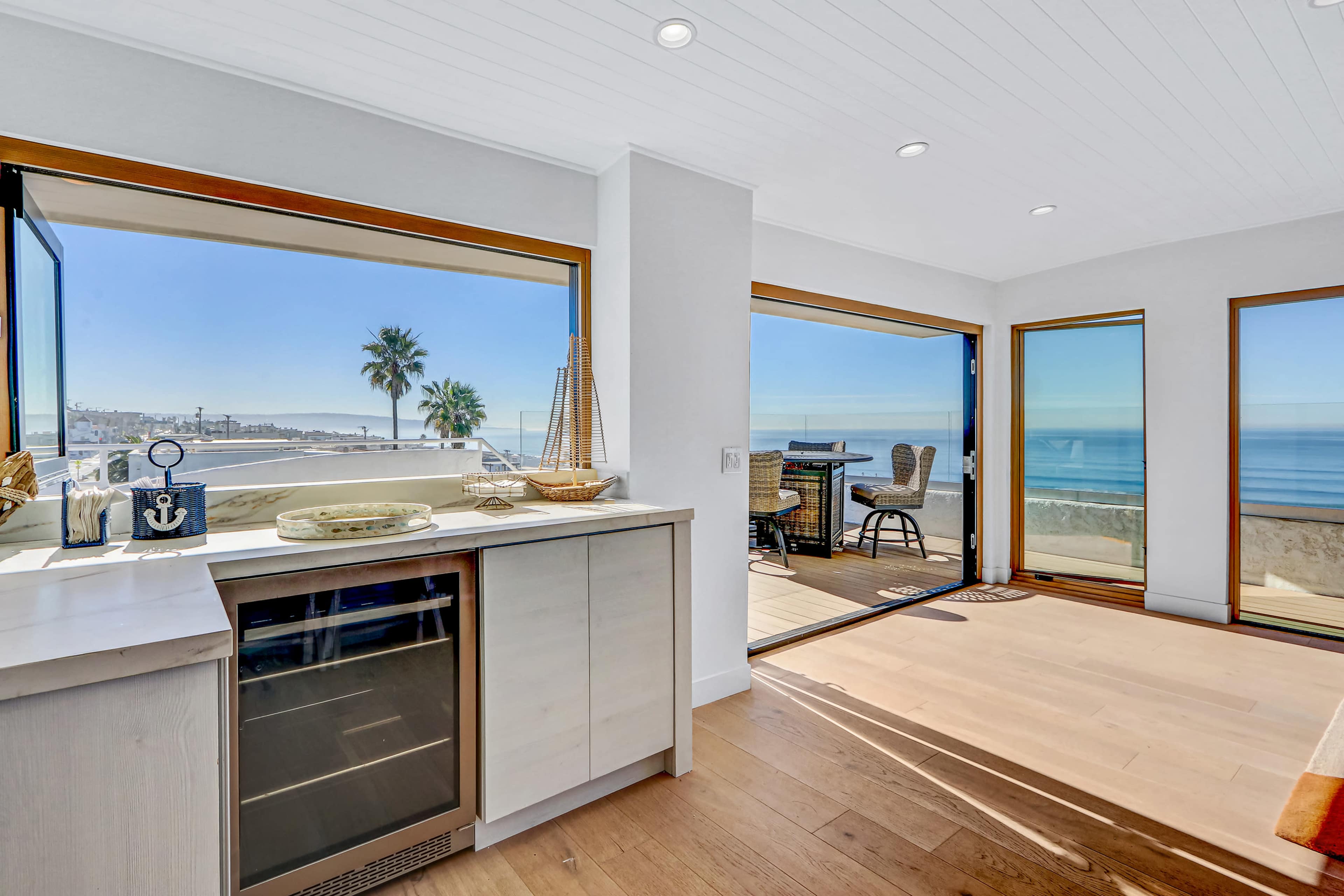 The image shows a modern coastal living space with large windows offering a view of the ocean and palm trees, featuring a bar area and outdoor seating.