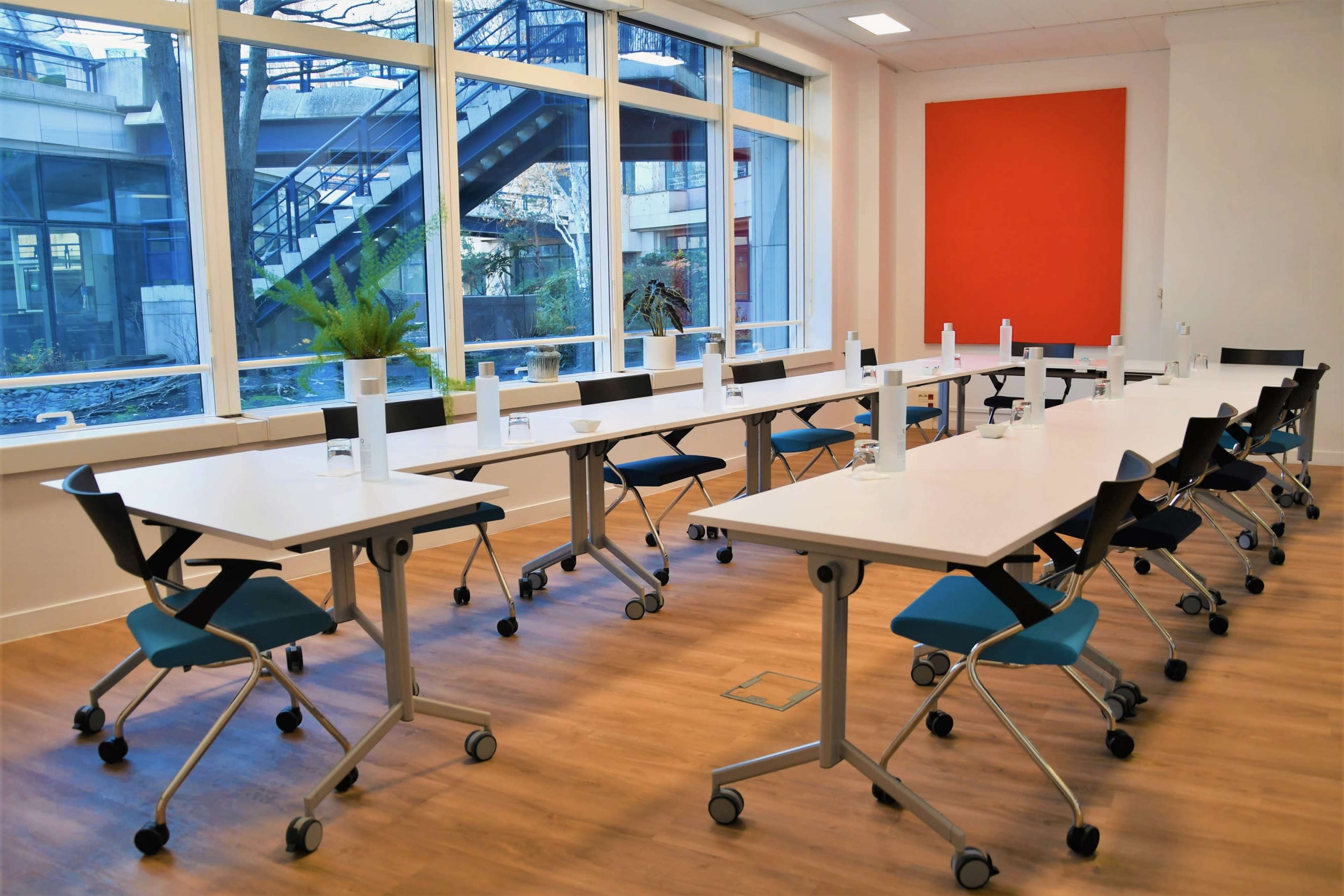 A spacious conference room features multiple movable tables arranged in an elongated shape, each equipped with a water bottle, and large windows providing natural light and a view of outdoor stairs and greenery.
