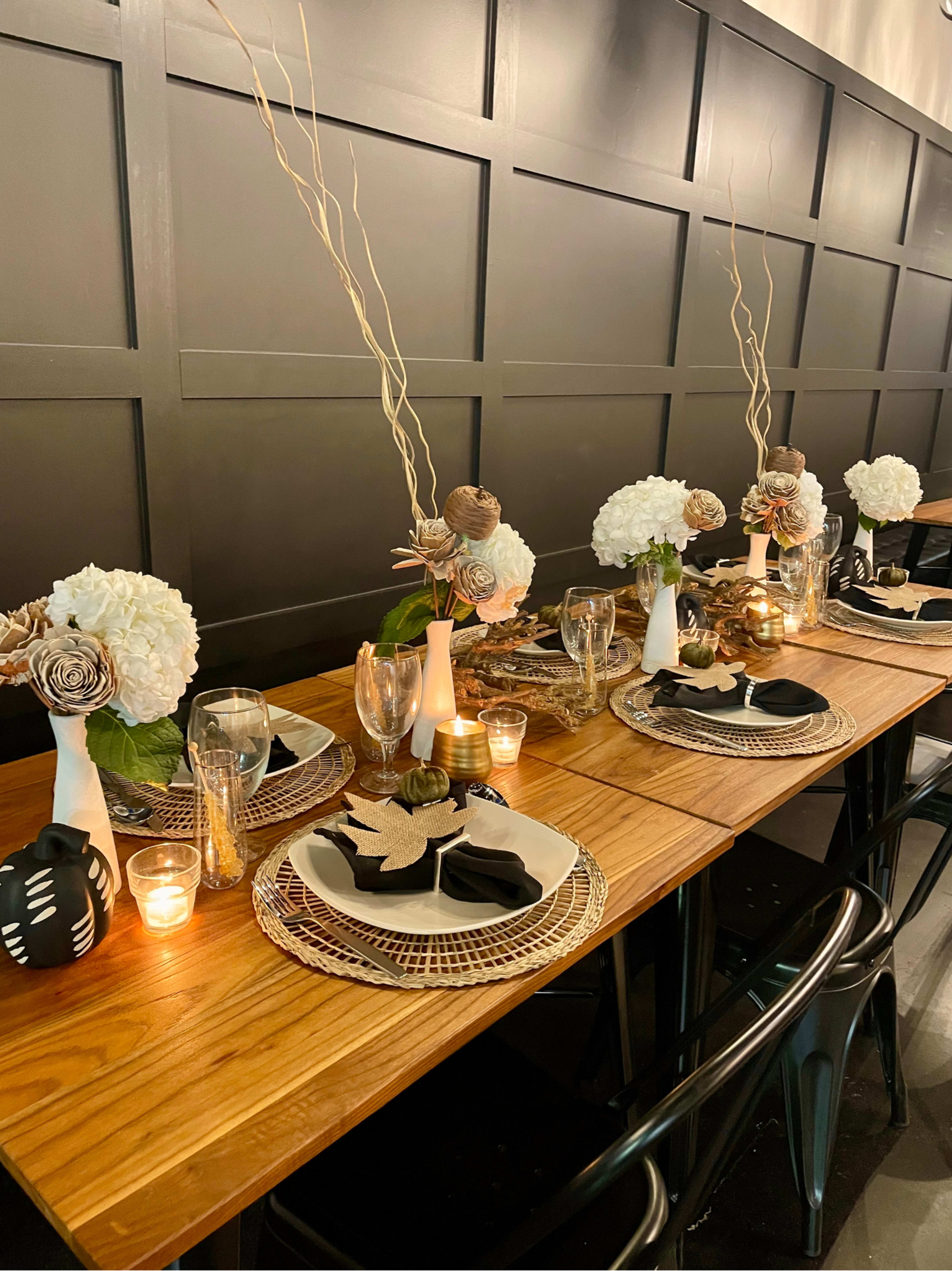 A long wooden dining table is elegantly set with plates, glasses, and decorative centerpieces featuring flowers and candles along a dark-paneled wall.