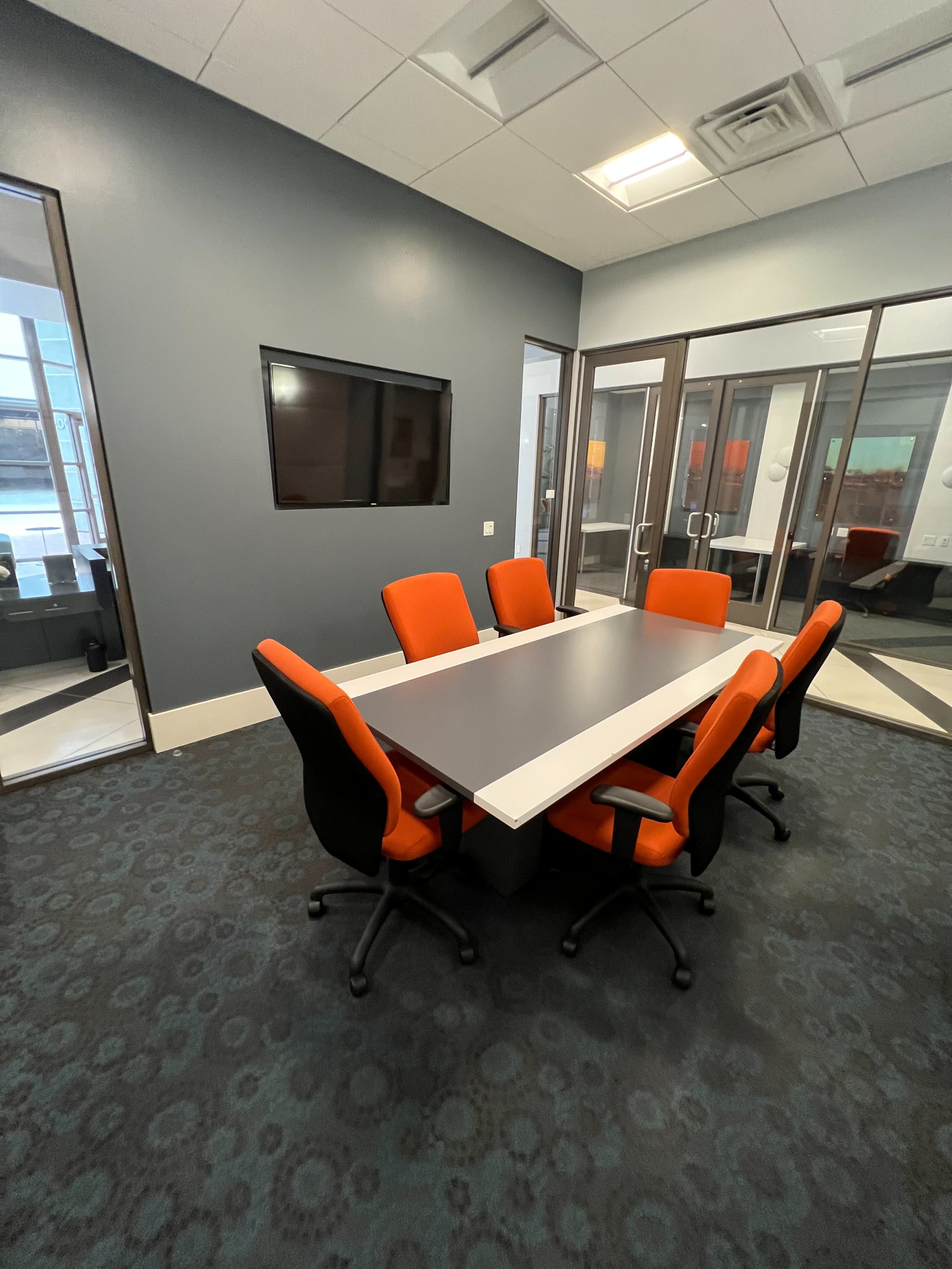 A modern conference room equipped with a rectangular table surrounded by six chairs, with a wall-mounted television and large windows in the background.