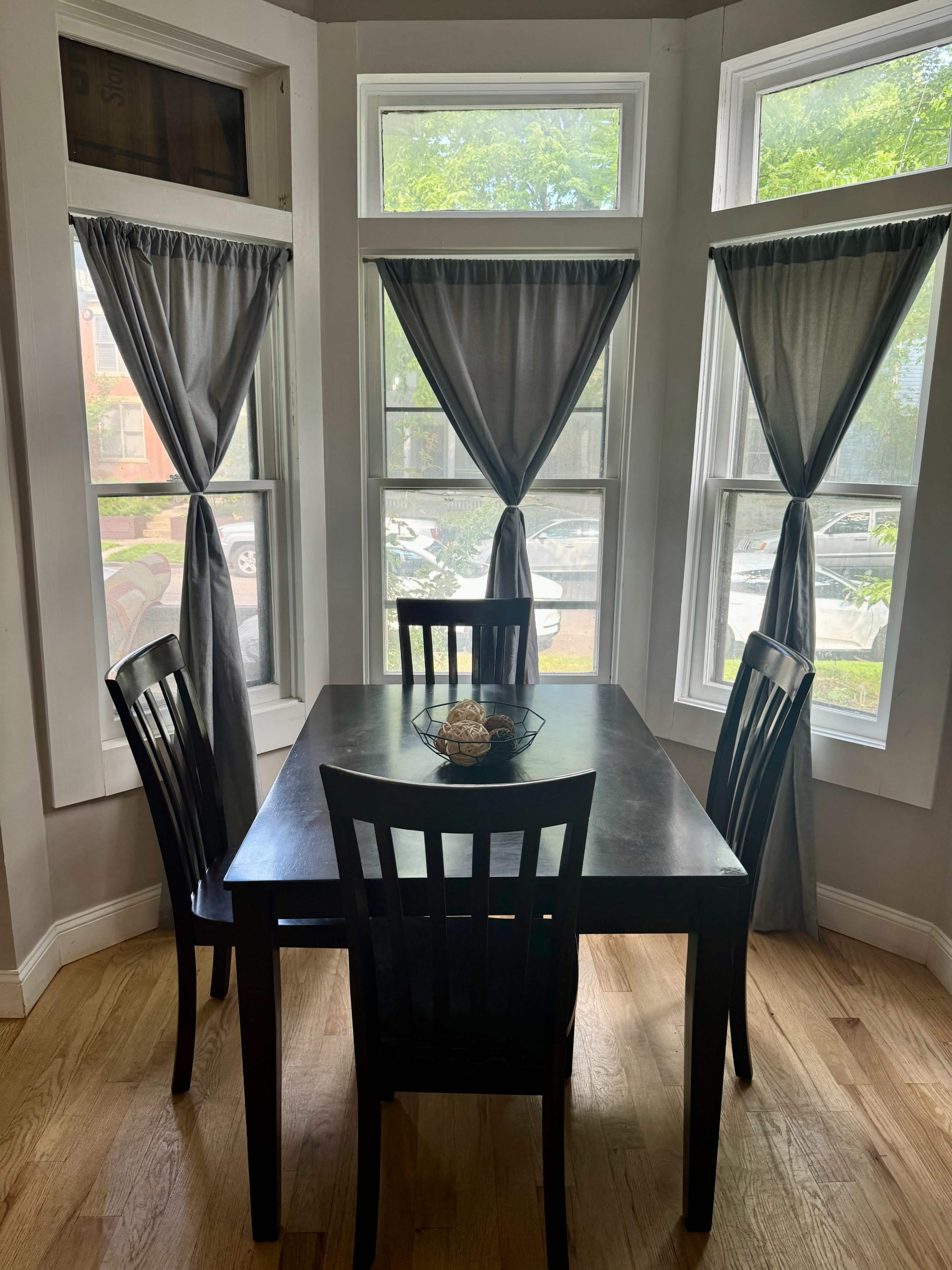A dark wooden table is set in a well-lit corner with four chairs and large windows dressed with gray curtains.