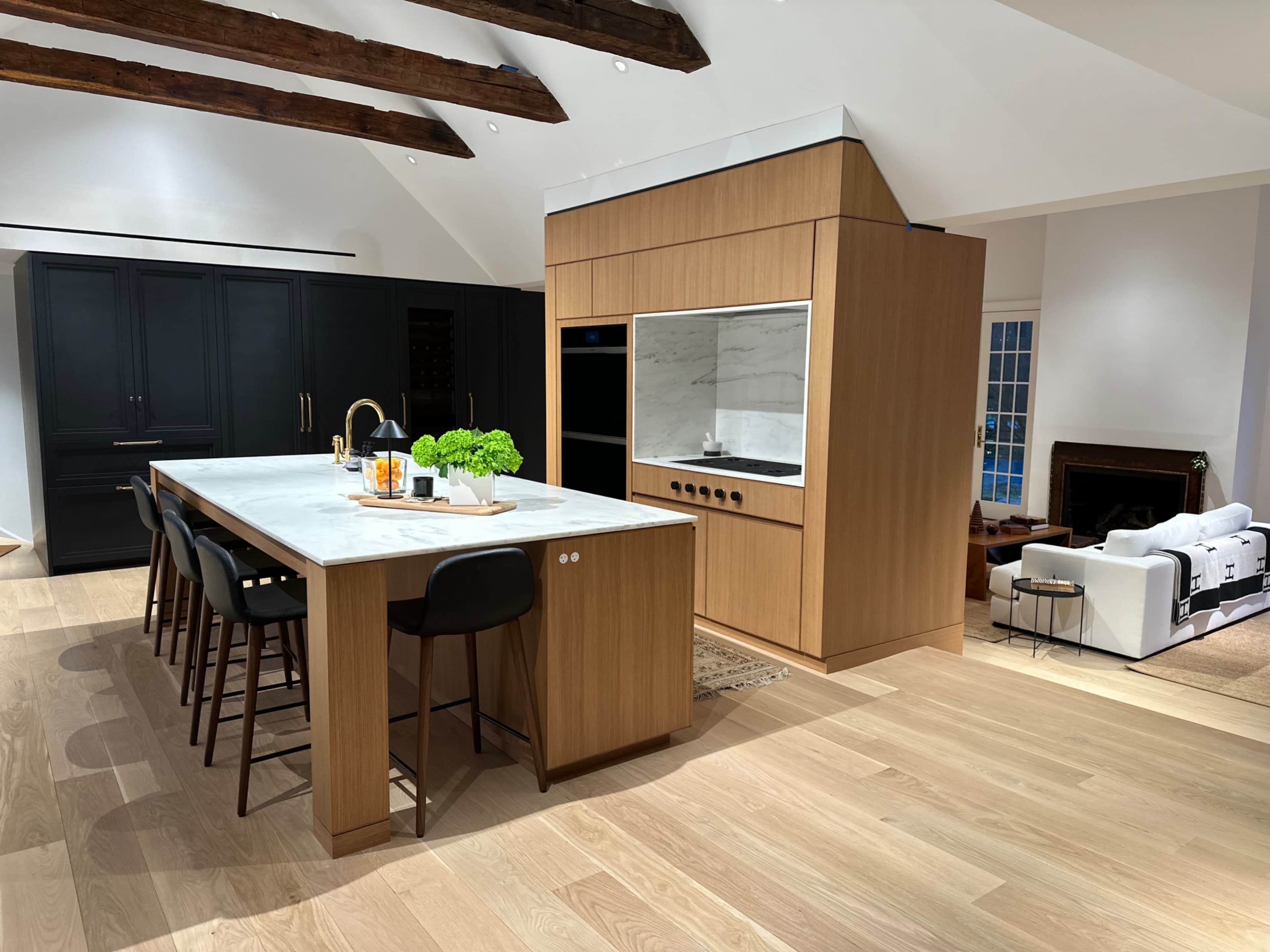 A modern kitchen features a large marble island with bar seating, wooden cabinetry, and a black accent wall, all set in an open living space.