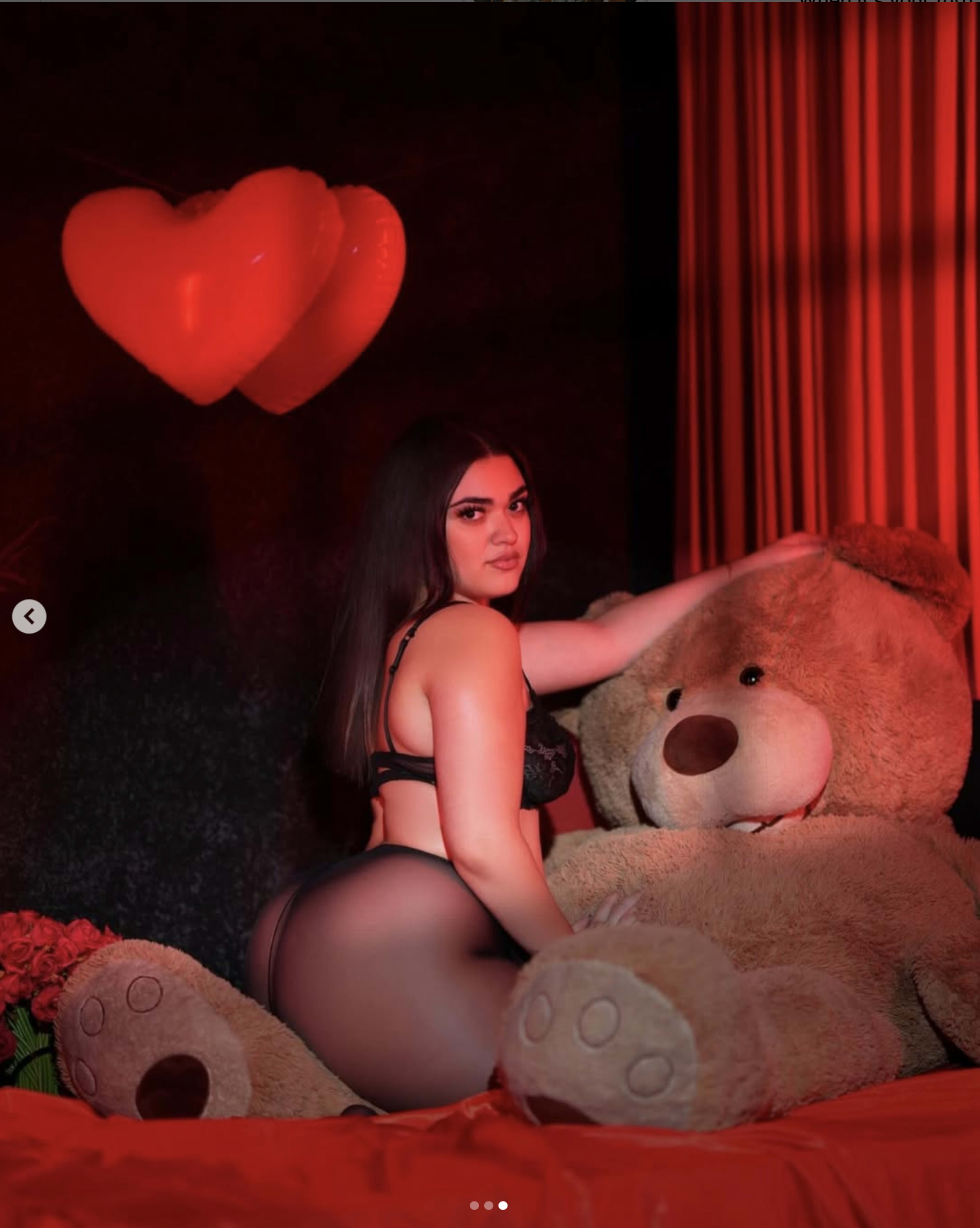 A woman in black lingerie poses beside a large teddy bear on a red-themed set with heart decorations.