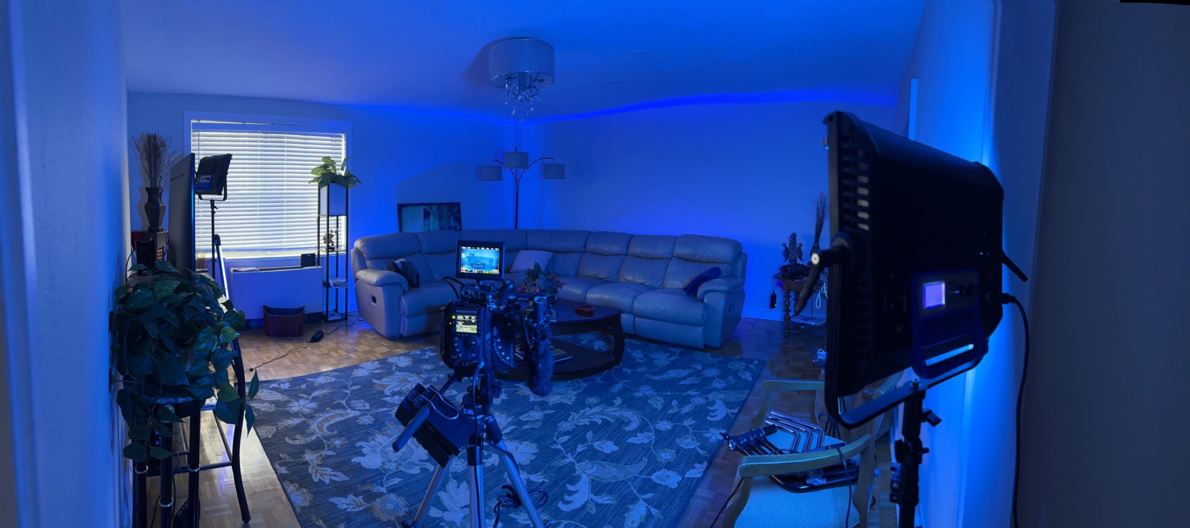 A living room is illuminated with blue lighting, featuring a sectional sofa, a large rug, and camera equipment set up for a shoot.