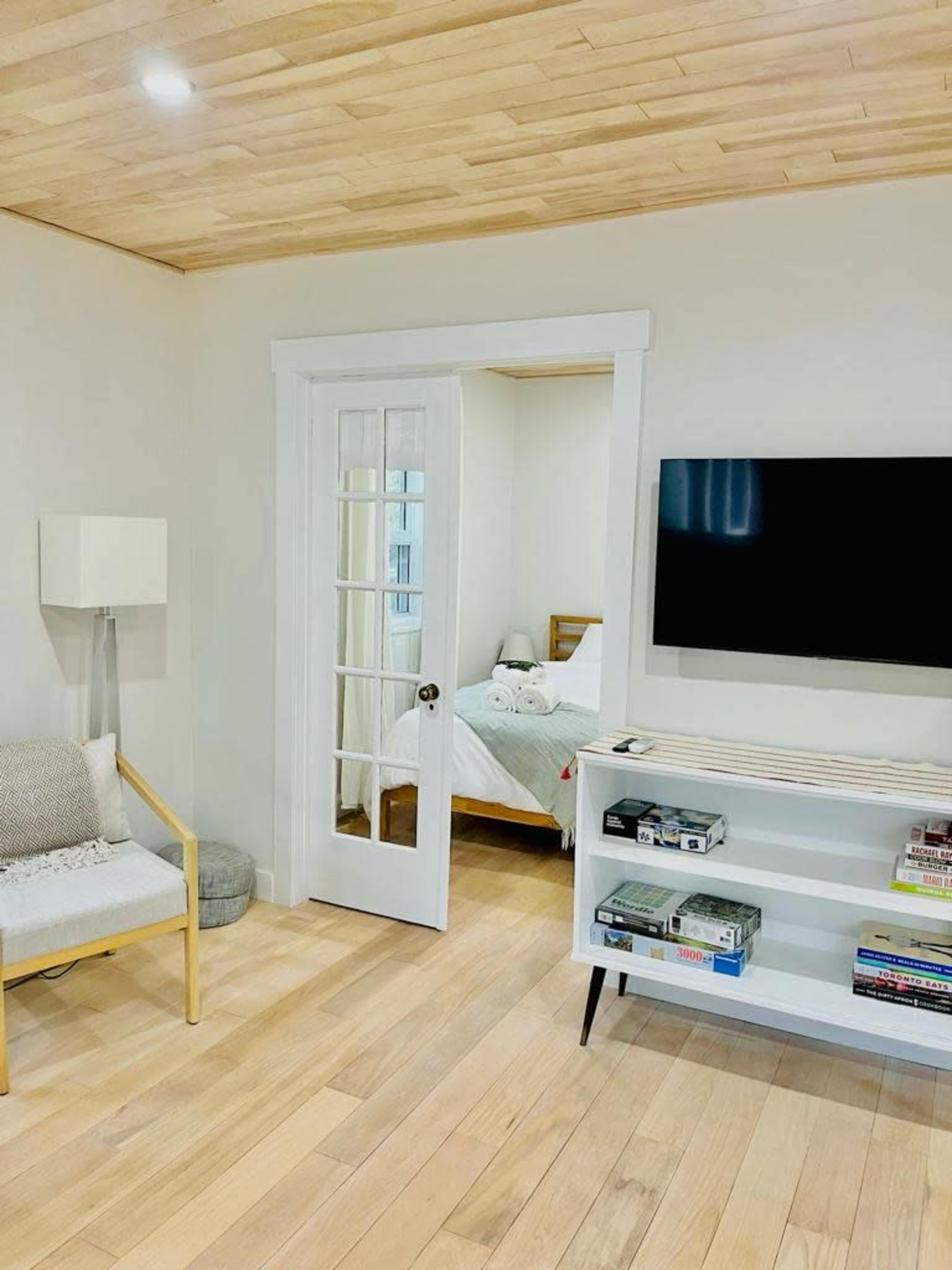 The image shows a modern living space with light wood flooring, a white bookshelf, a television mounted on the wall, and a door leading to a bedroom.