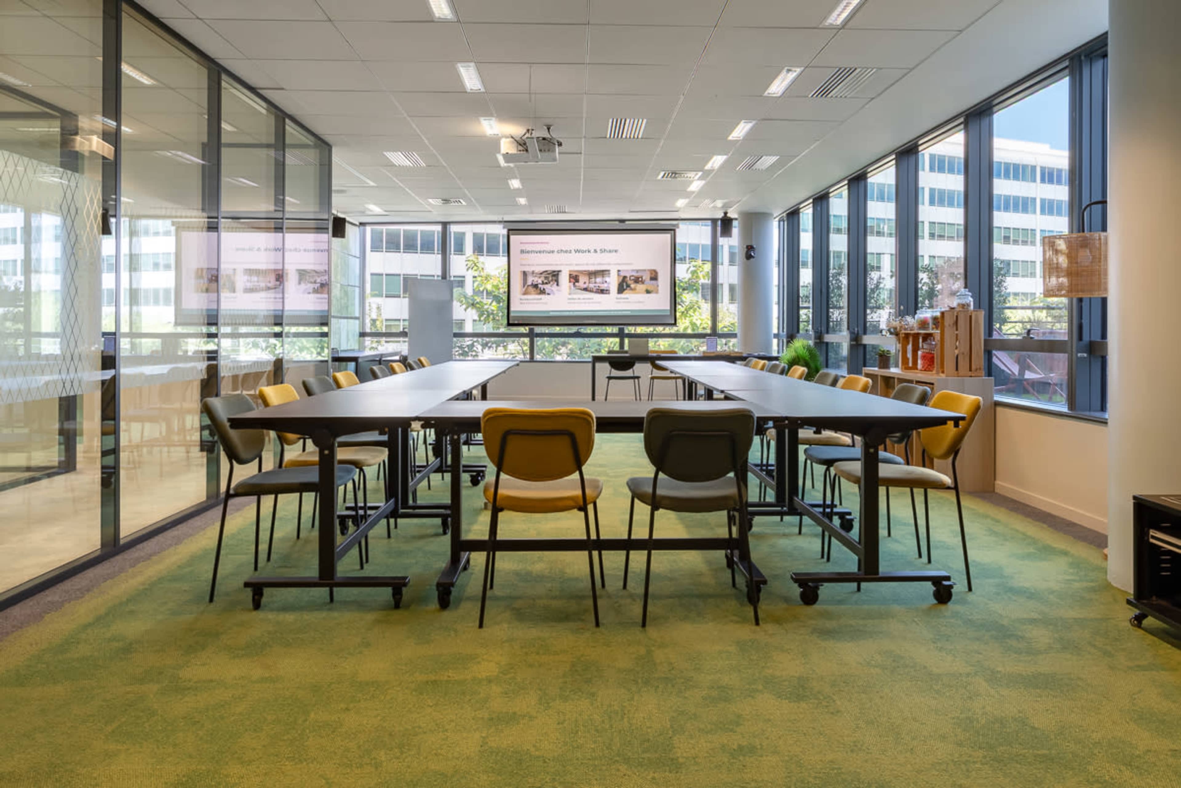 The image shows a modern conference room with a large rectangular table, rolling chairs, and large windows displaying an urban landscape.