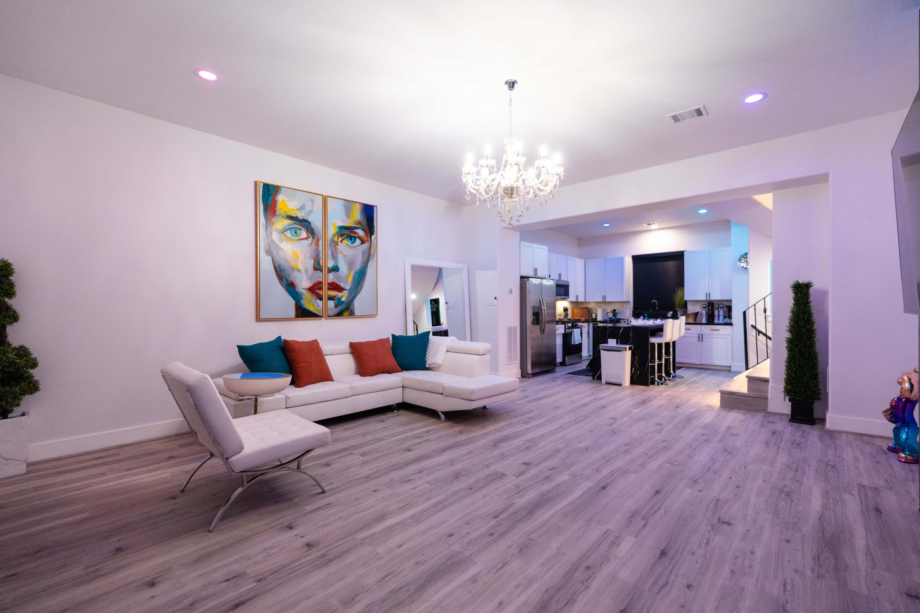 A modern living room features a white sectional sofa with colorful cushions, a large artwork on the wall, and an open kitchen area with bar seating.