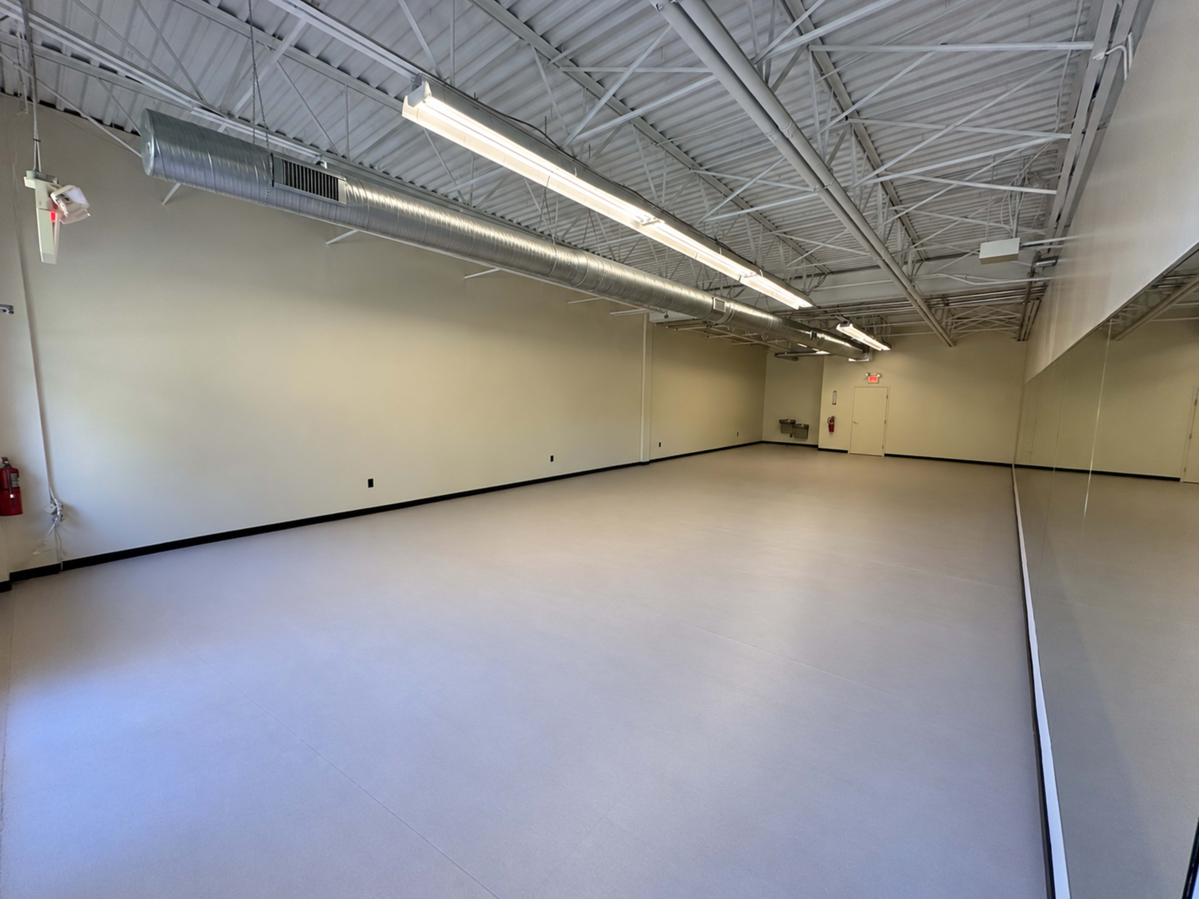 The image shows a large, empty room with a smooth, light-colored floor and a wall of mirrors, illuminated by overhead fluorescent lights.