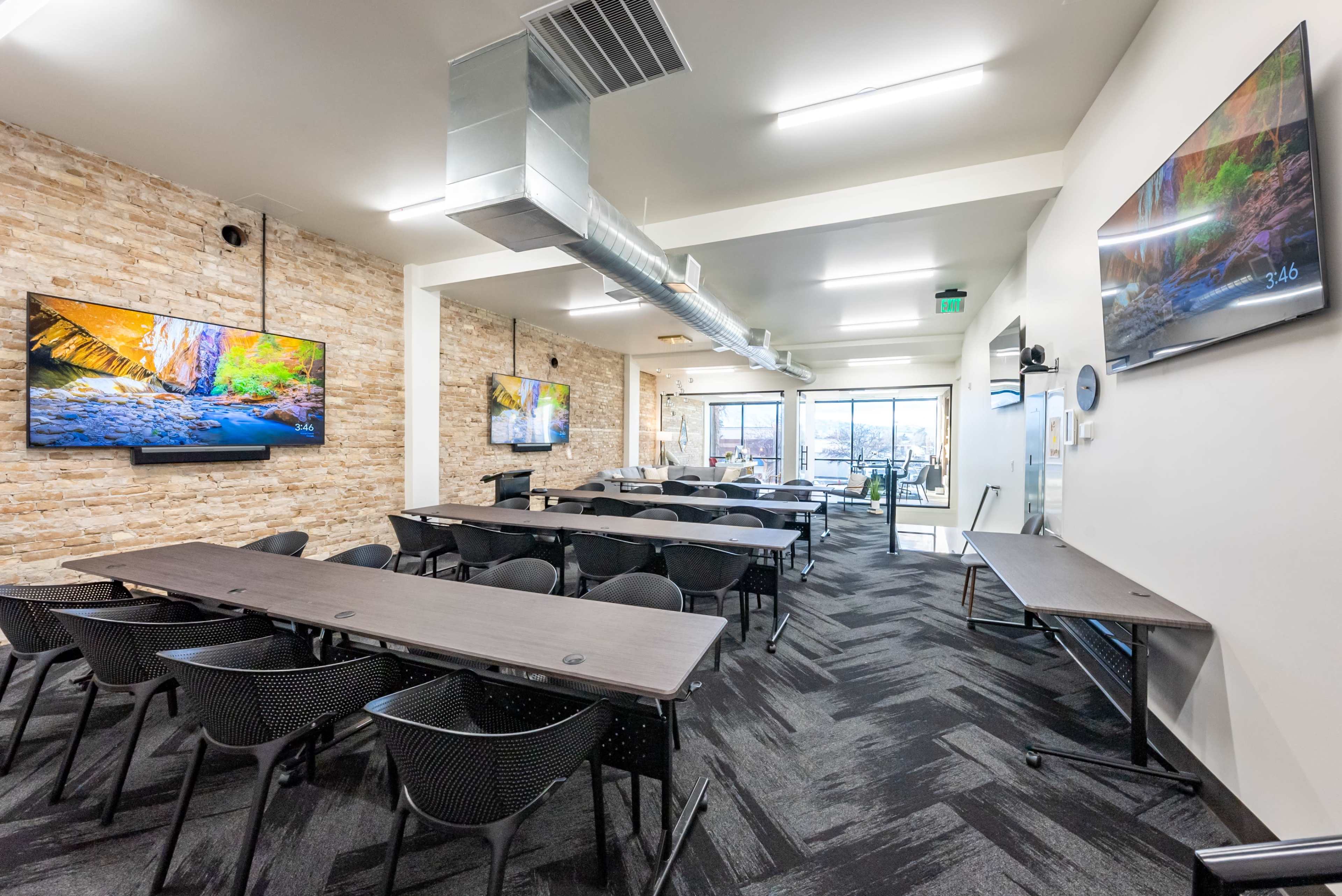 A modern meeting room with two large screens mounted on the walls, long tables arranged for participants, and an open view to an external area through glass doors.