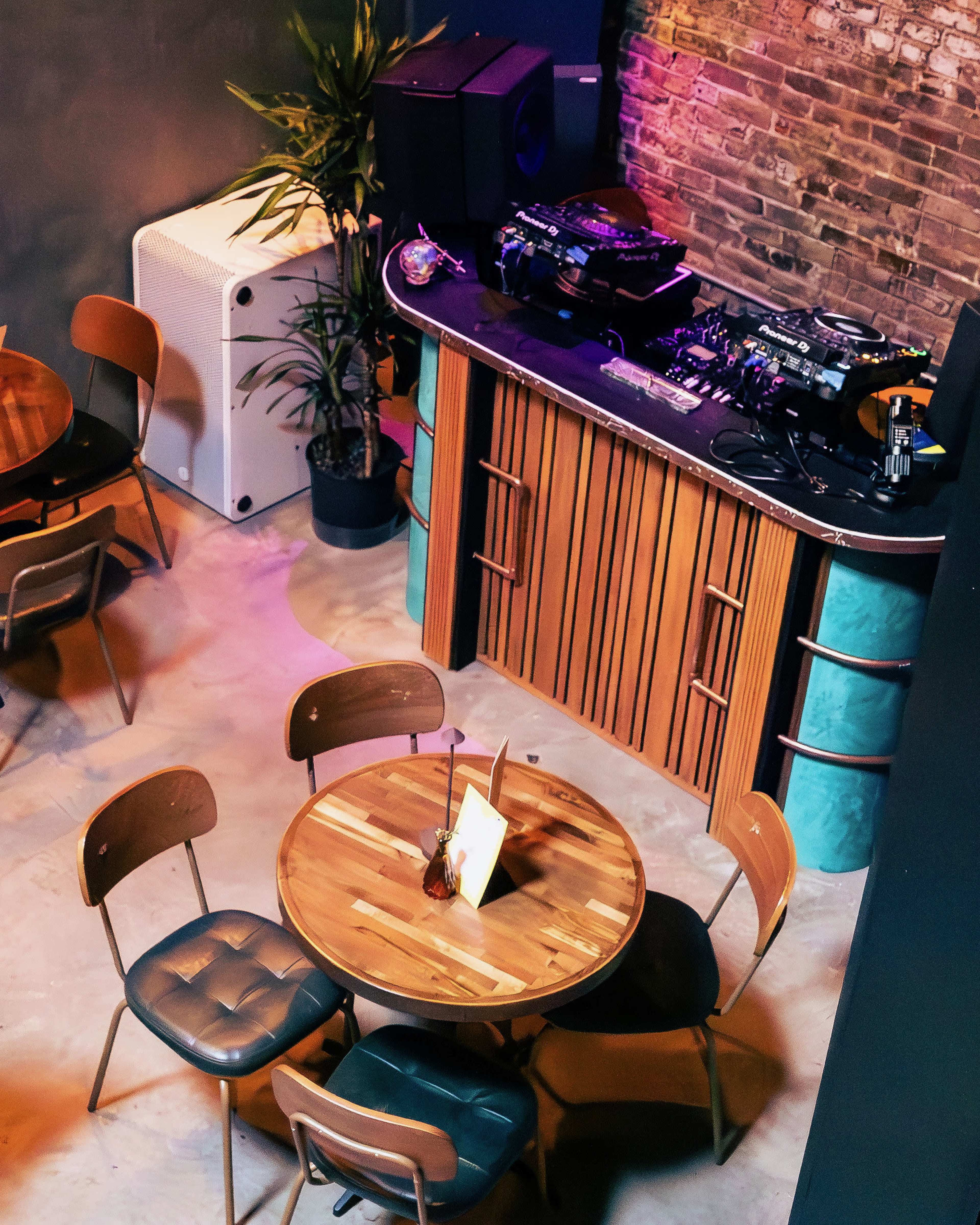 The image shows a cozy interior of a venue featuring wooden tables and chairs, a DJ setup, and exposed brick walls.