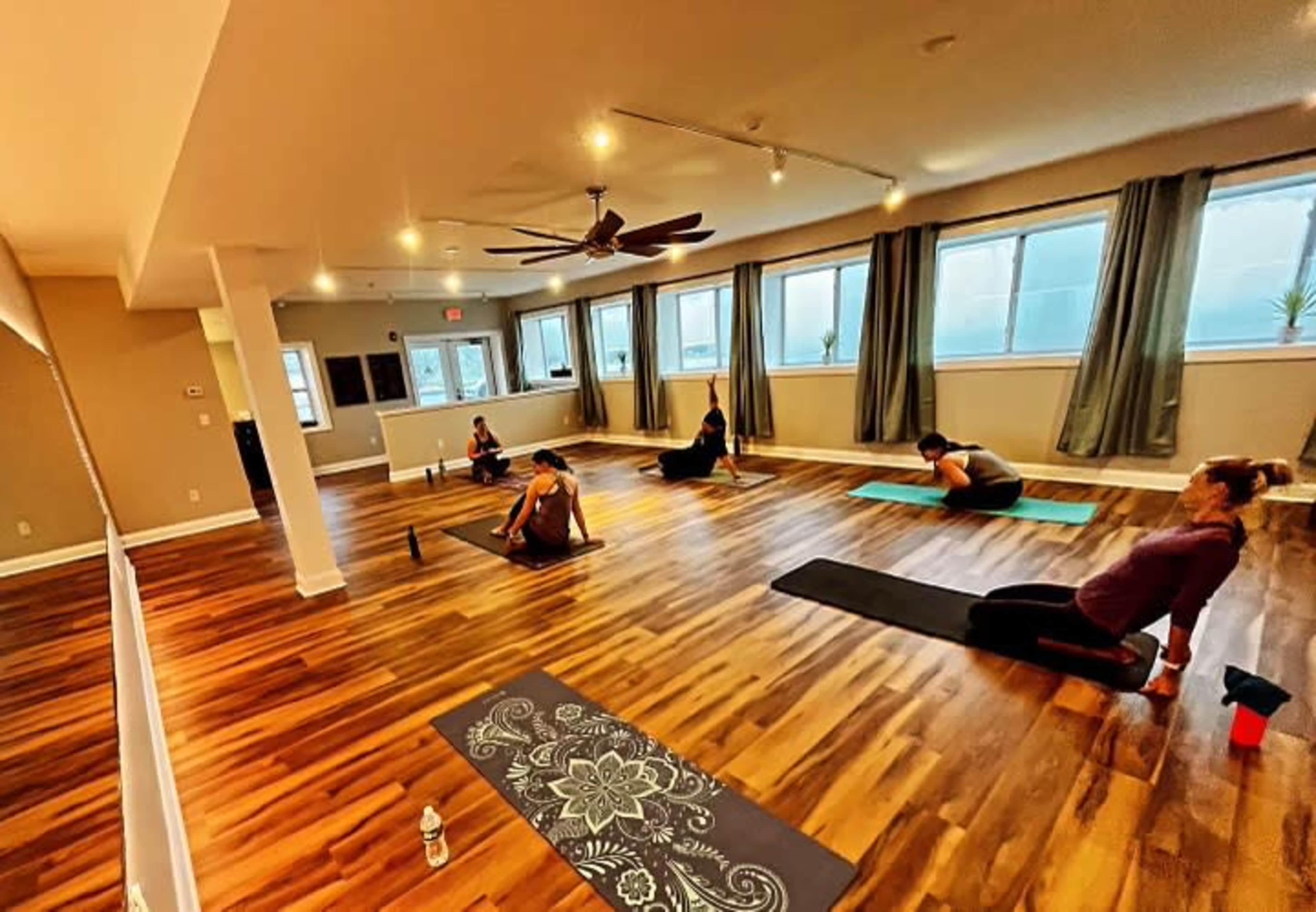 A group of individuals practices yoga on mats in a brightly lit studio with large windows and wooden flooring.