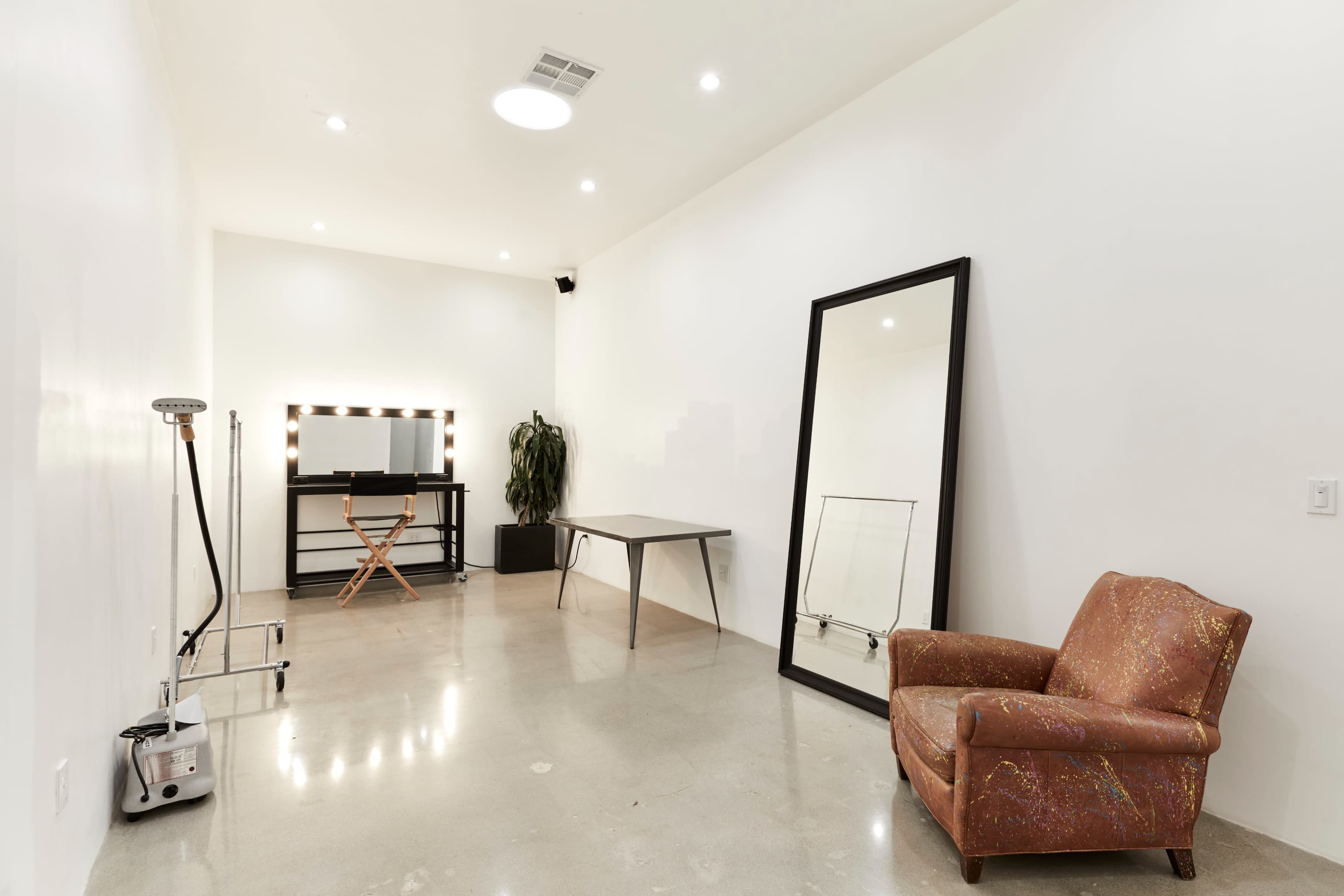 The image shows a minimalist room with polished concrete floors, a large mirror, a table, a chair, and a makeup station with lights.