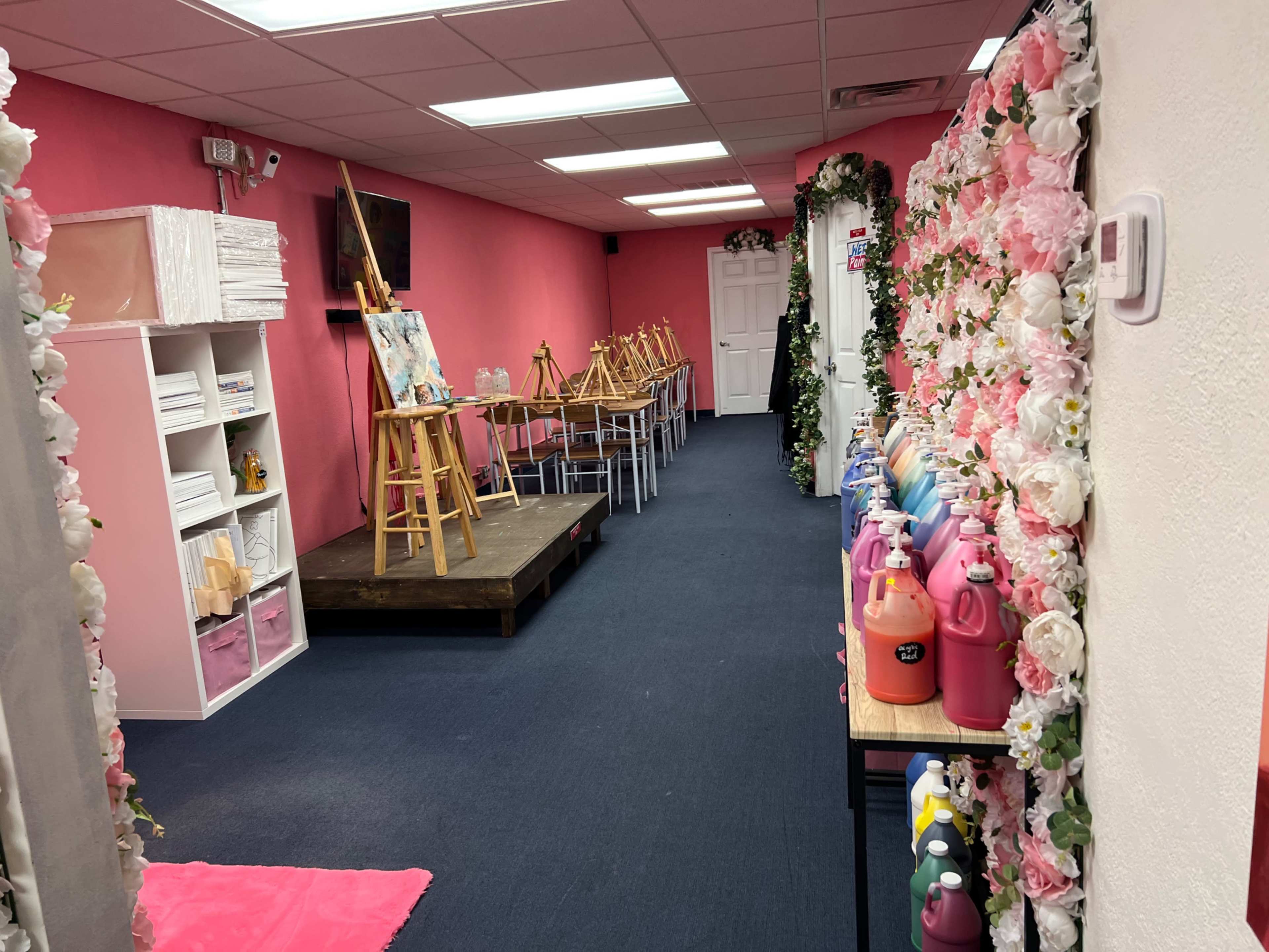 The image shows a brightly colored art studio with pink walls, a small stage for painting, easels set up, and neatly arranged bottles of paint along one side.
