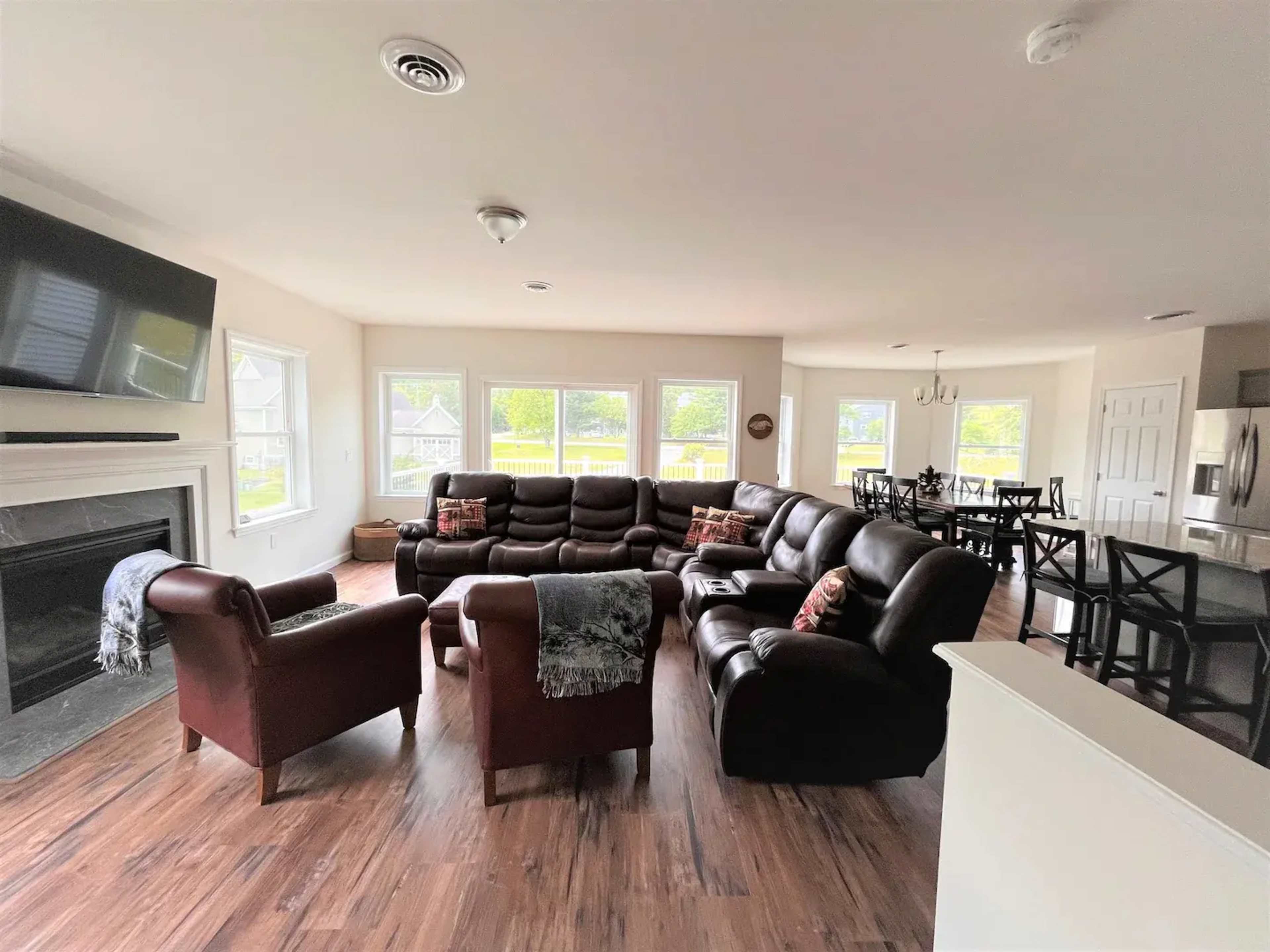 The room features a seating area with brown leather couches and armchairs arranged around a large TV, with windows showing a view of the outdoors.
