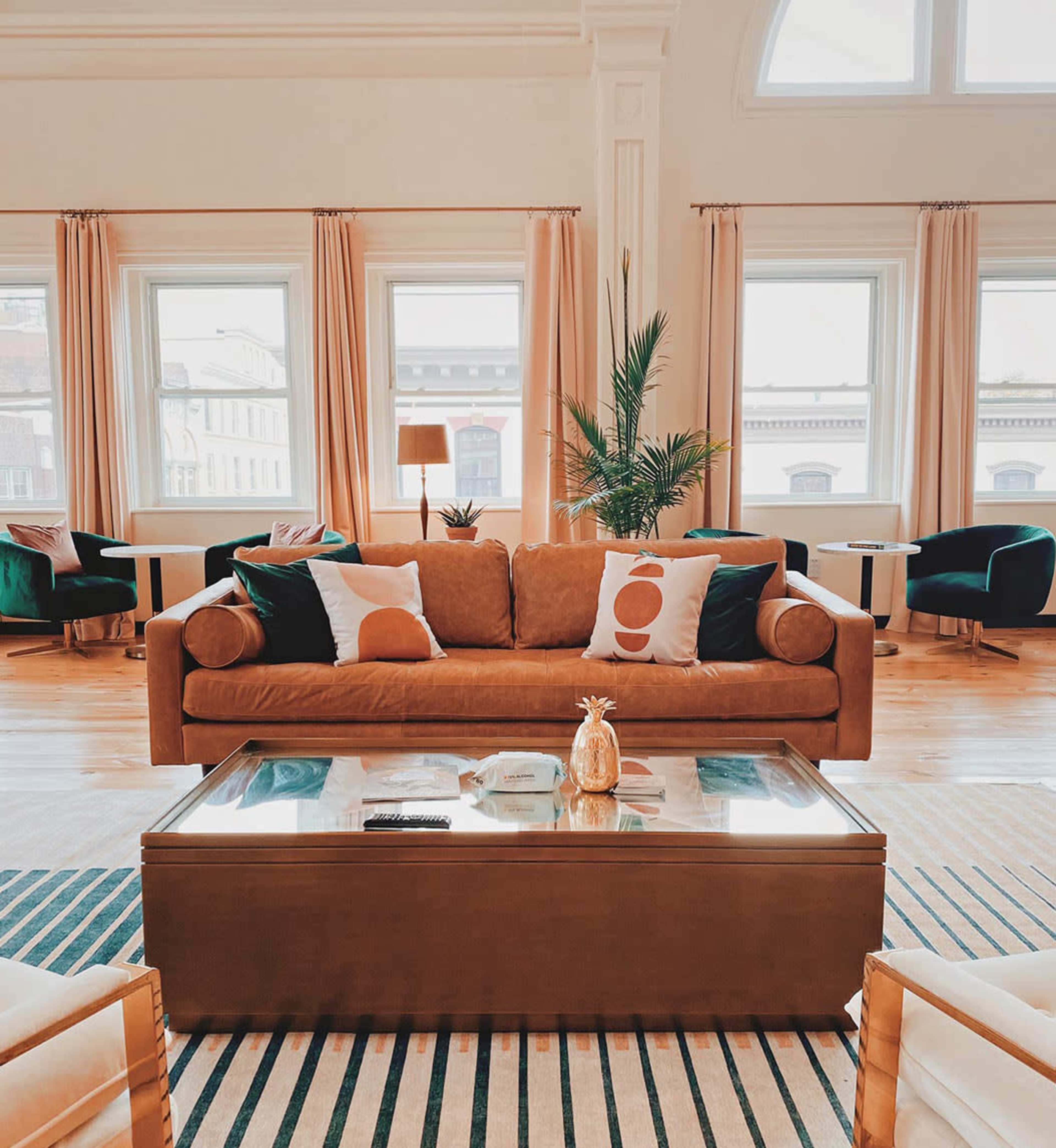 A spacious living room features a brown sofa with decorative pillows, a glass-topped coffee table, and several chairs by large windows.
