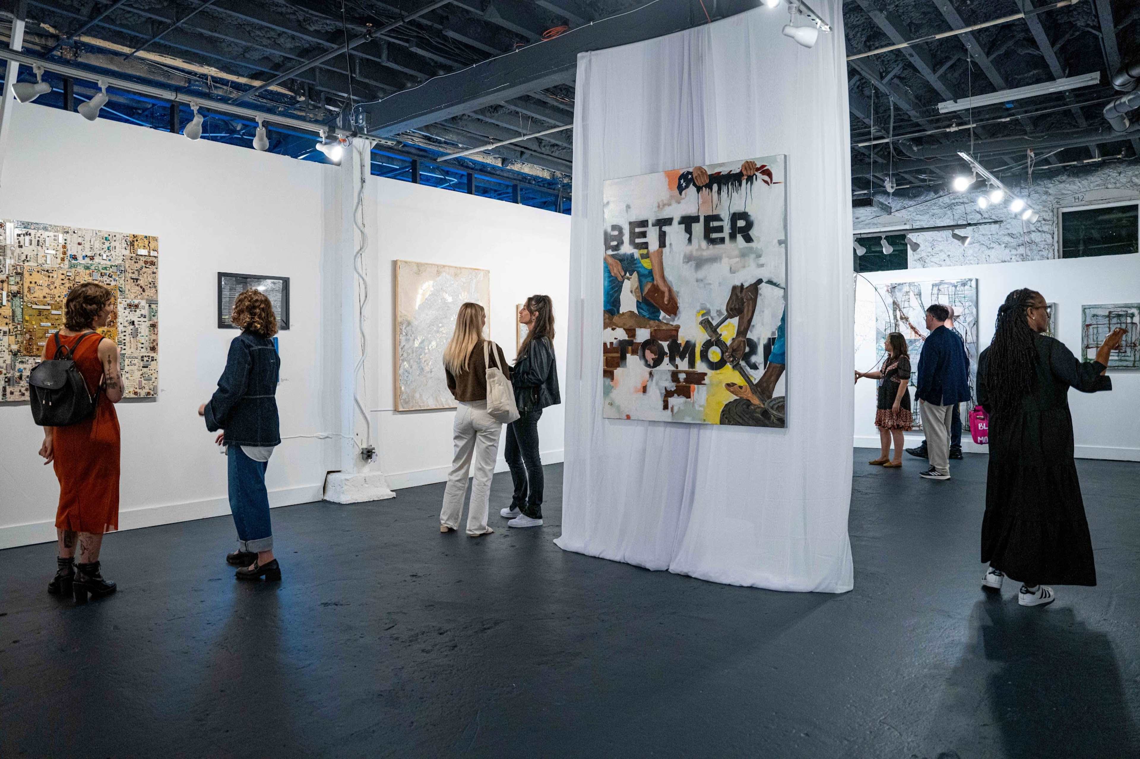 Several people are viewing contemporary artworks in a well-lit gallery space, with various paintings displayed on white walls.