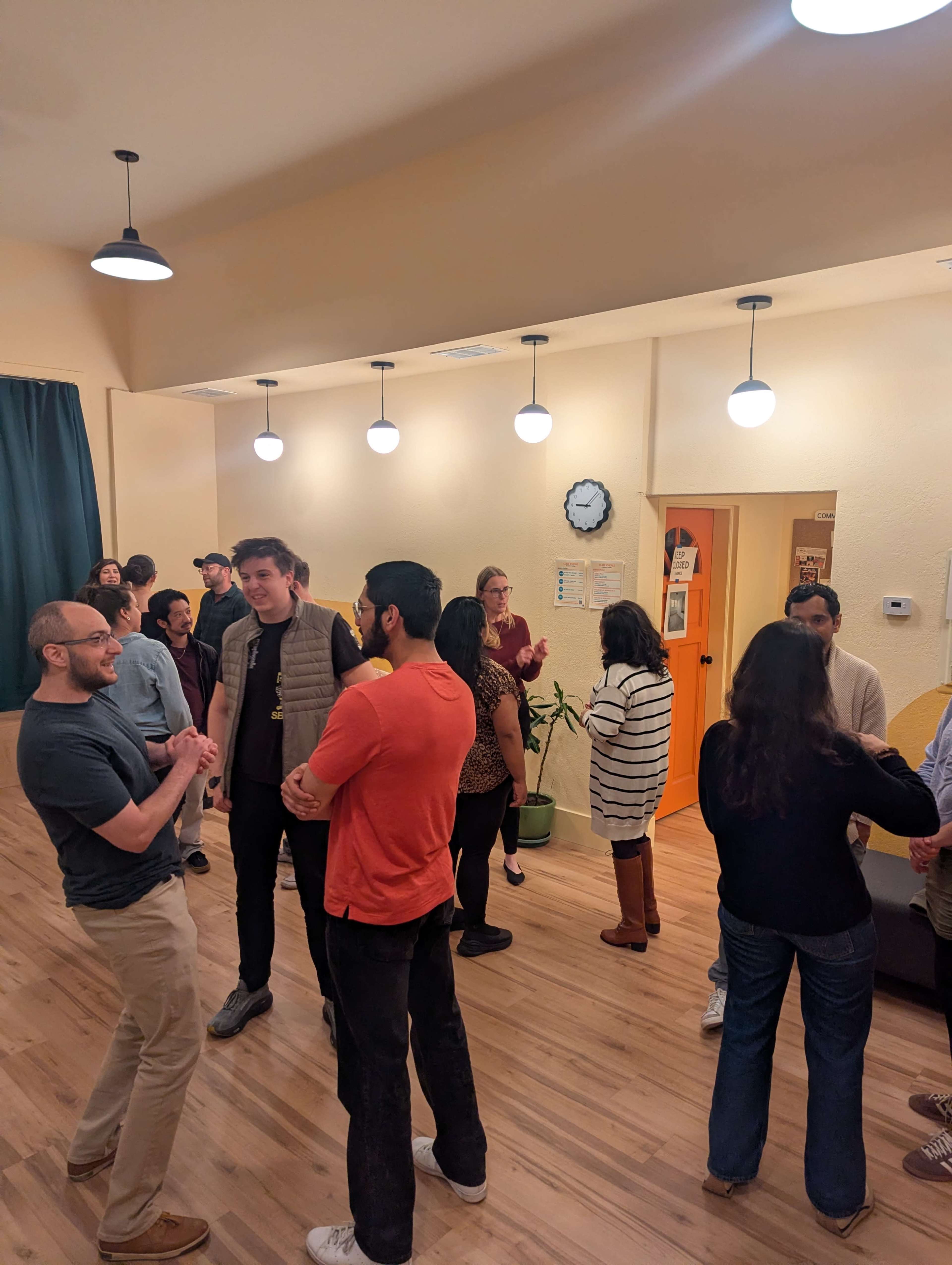 A group of people engages in conversation in a spacious, brightly lit interior with a wooden floor and plants.