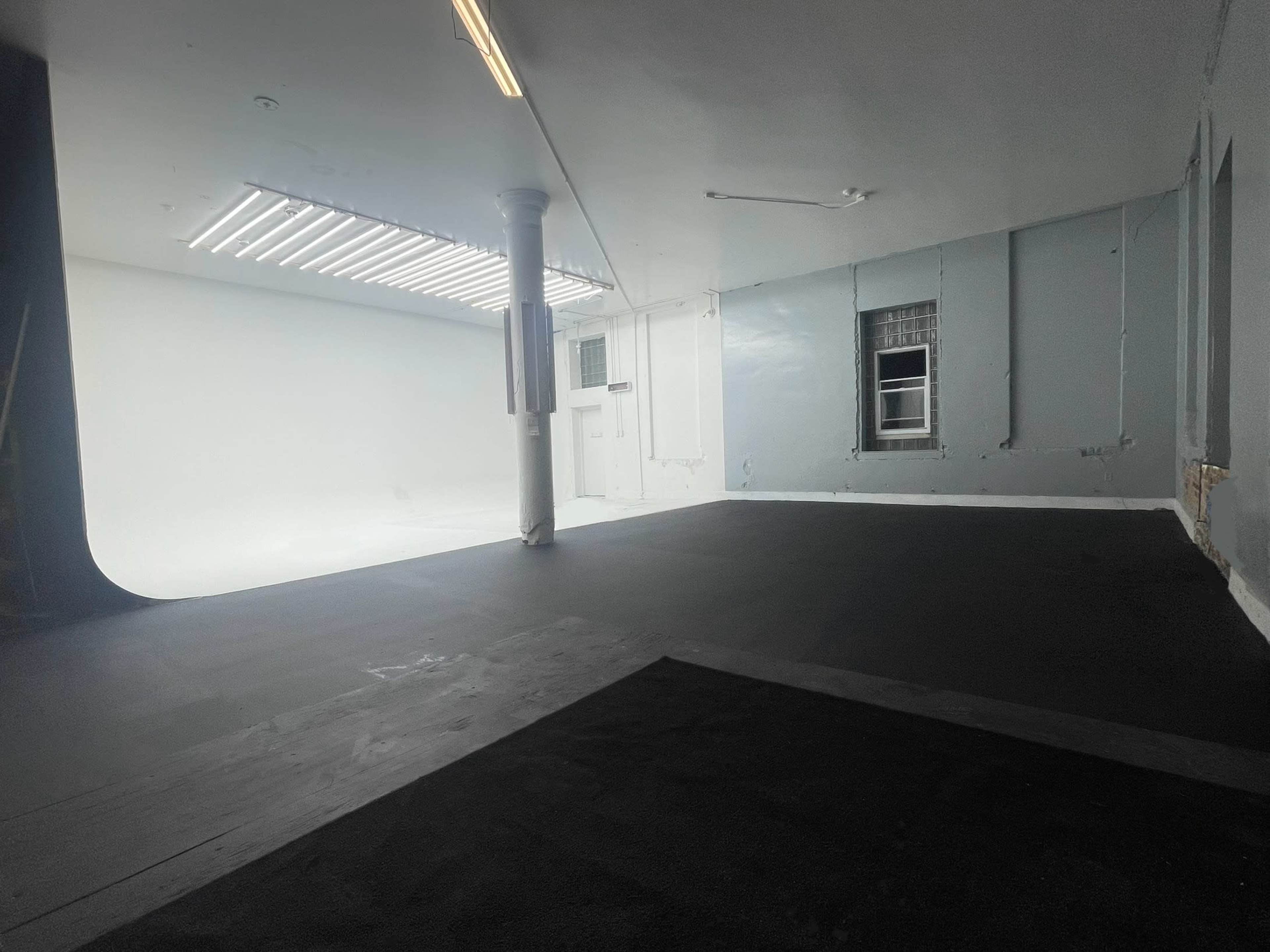 A spacious studio with black flooring, white walls, and a large open area illuminated by fluorescent lights.