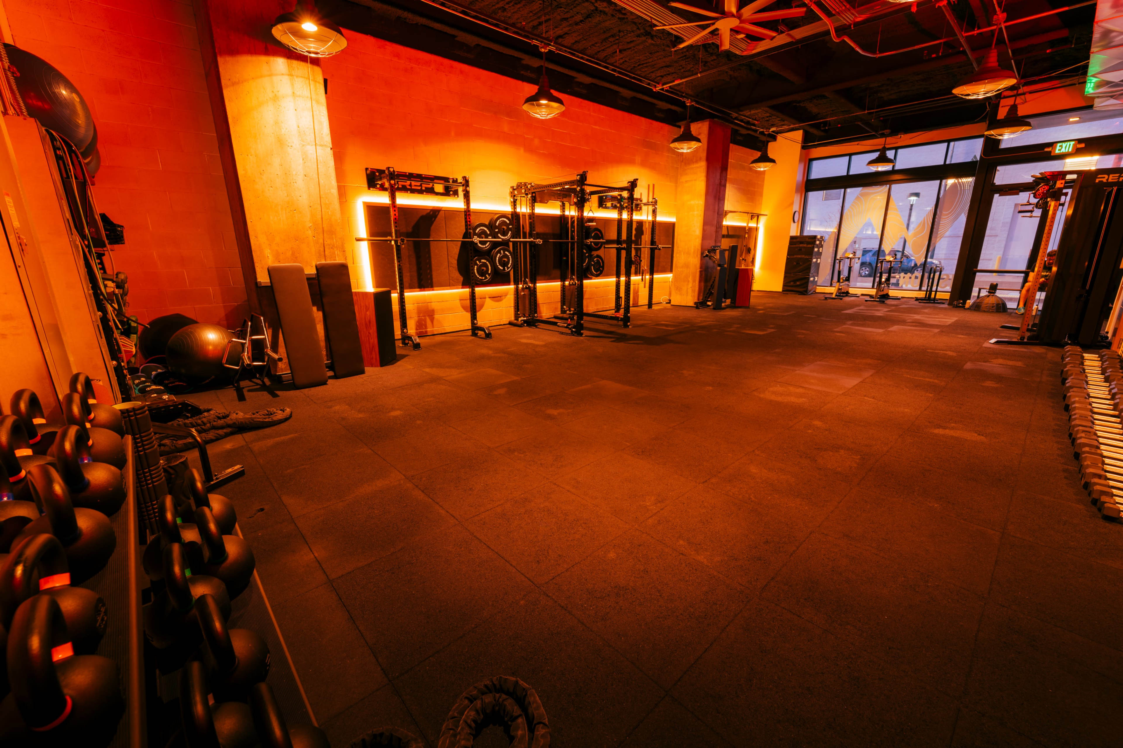 The image shows a spacious gym interior with orange lighting, featuring various workout equipment and an expansive window with a view outside.