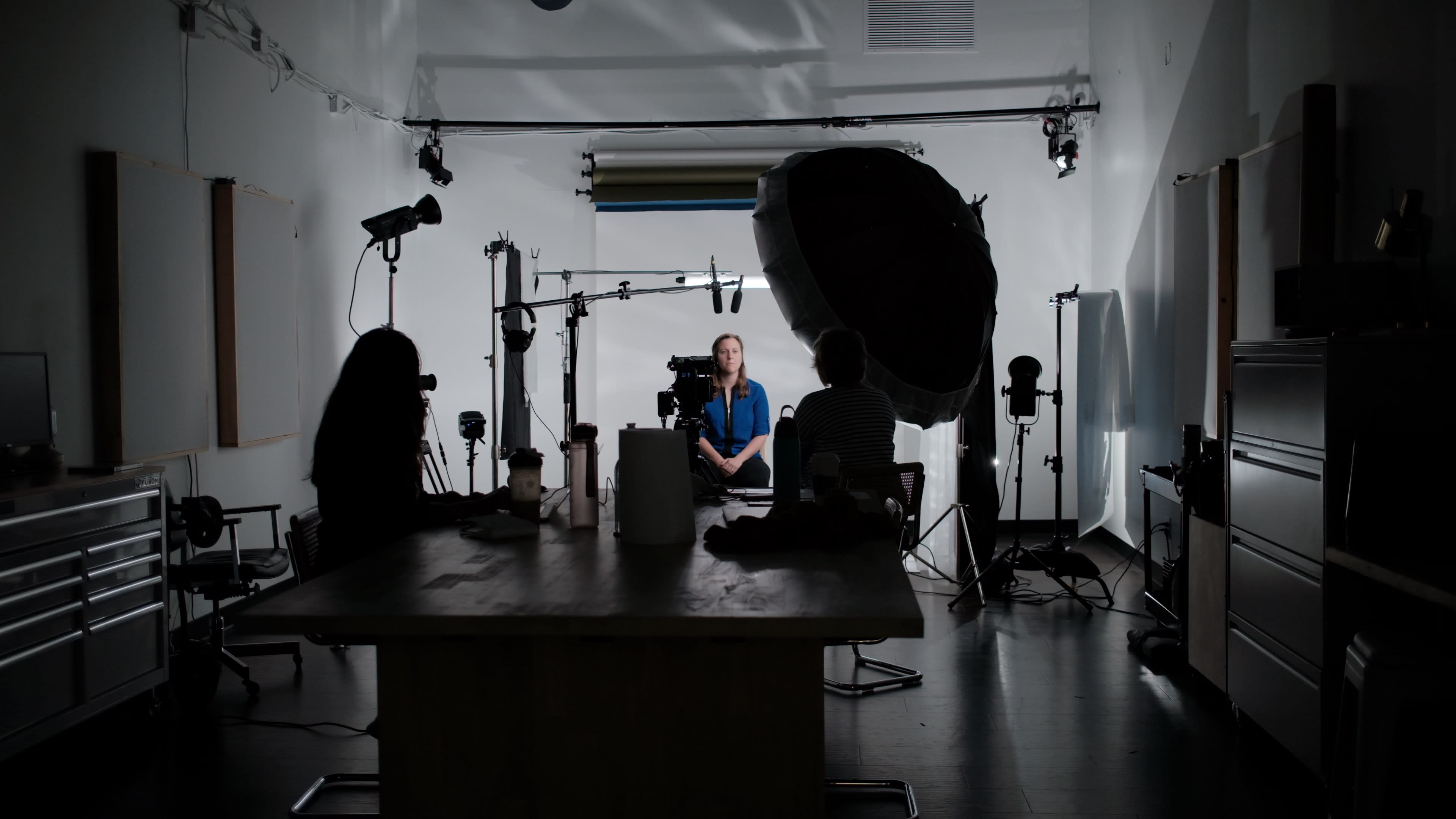 The image shows a well-lit interview setup in a studio, with a subject seated in the center and camera equipment positioned around.