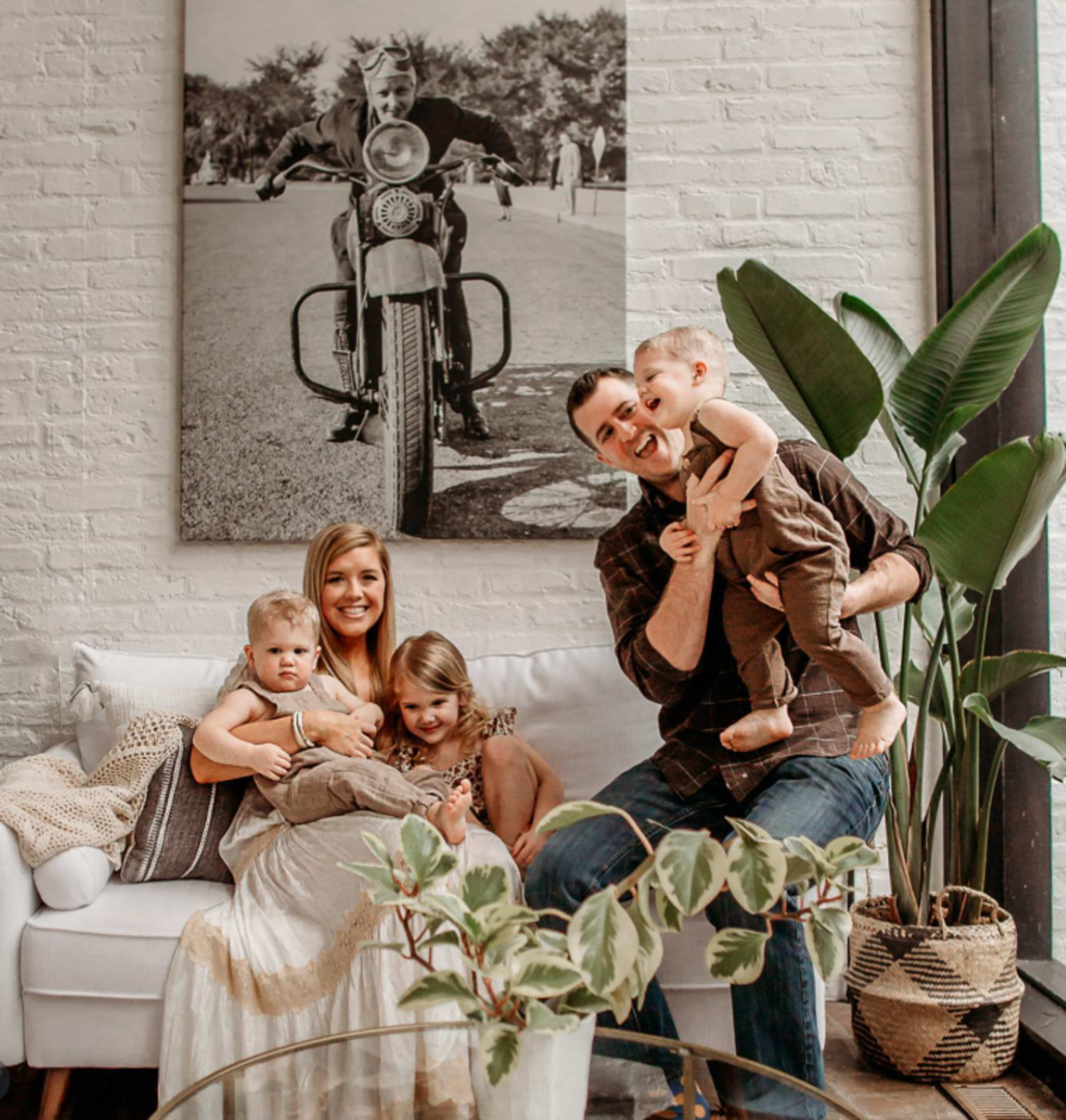A family of five sits together on a couch in a modern living room, with a large black-and-white photograph of a man on a motorcycle displayed on the wall behind them.