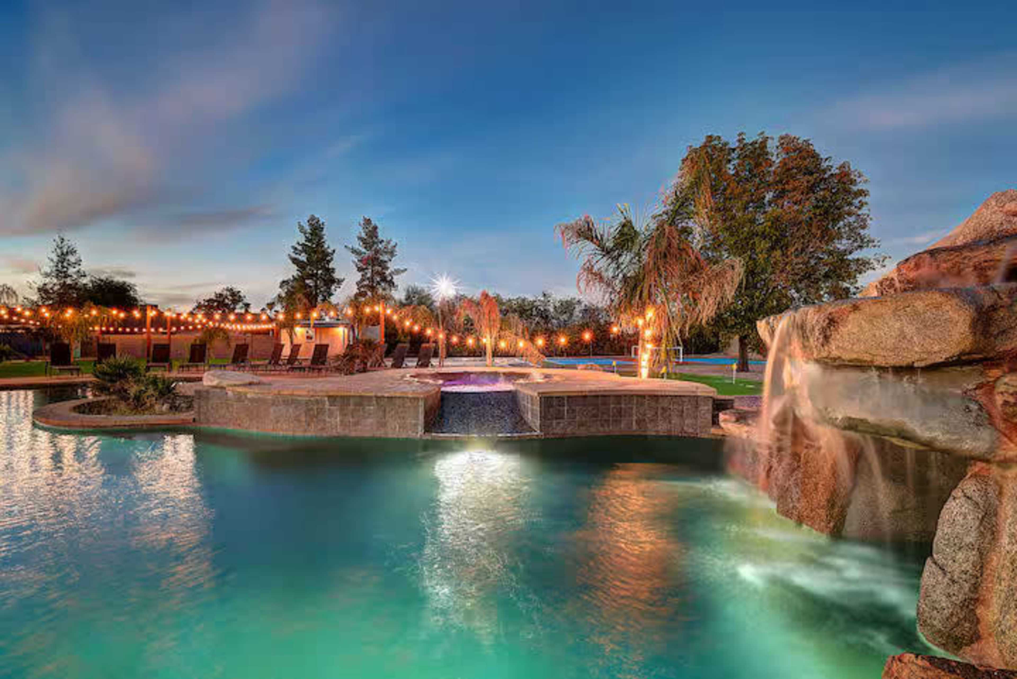The image shows a landscaped pool area featuring a waterfall, tropical plants, and string lights in the evening sky.