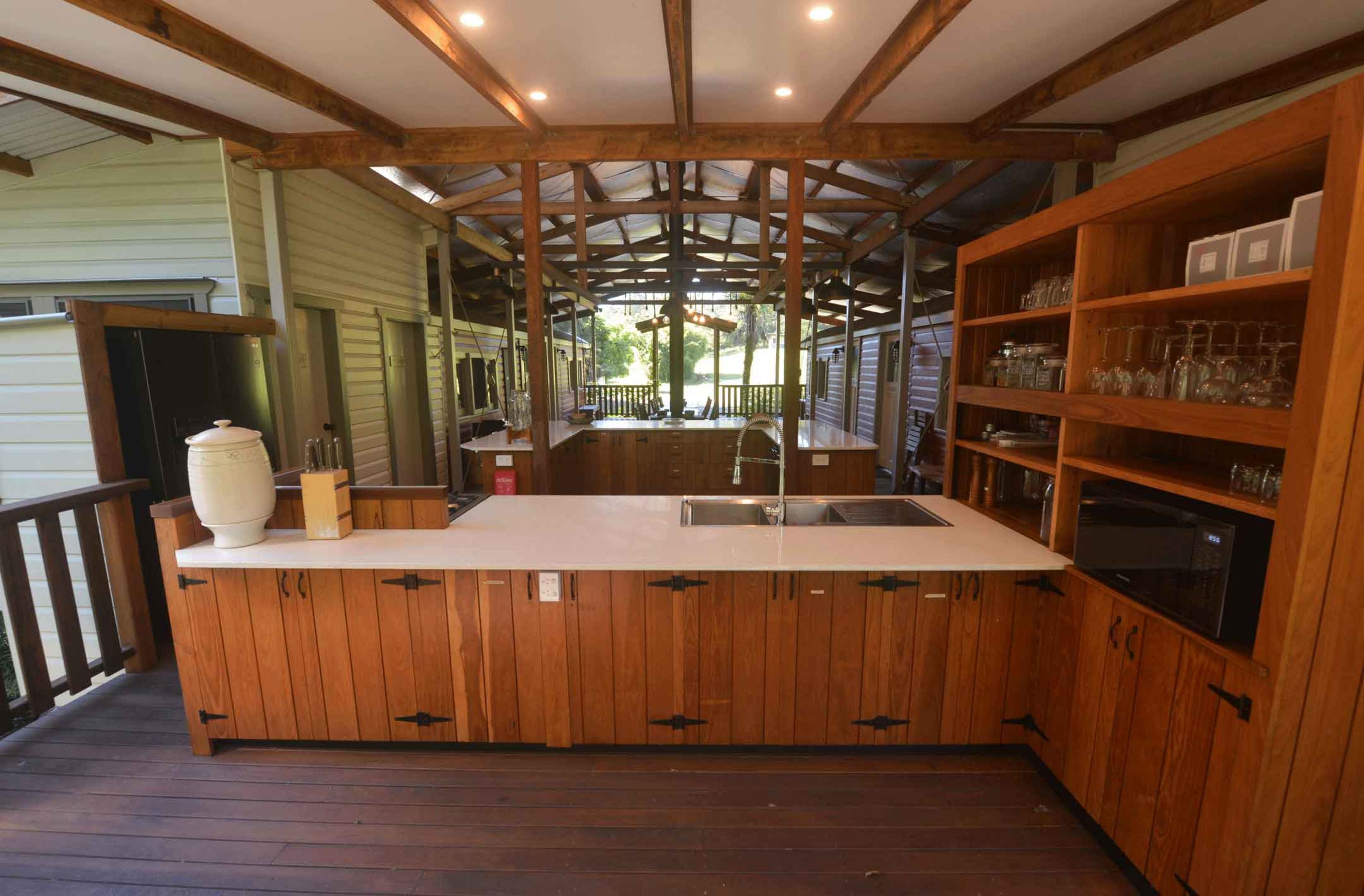 The image shows a wooden kitchen with a central island, featuring a double sink, a microwave, and shelves containing glassware, all set in an open, covered space.