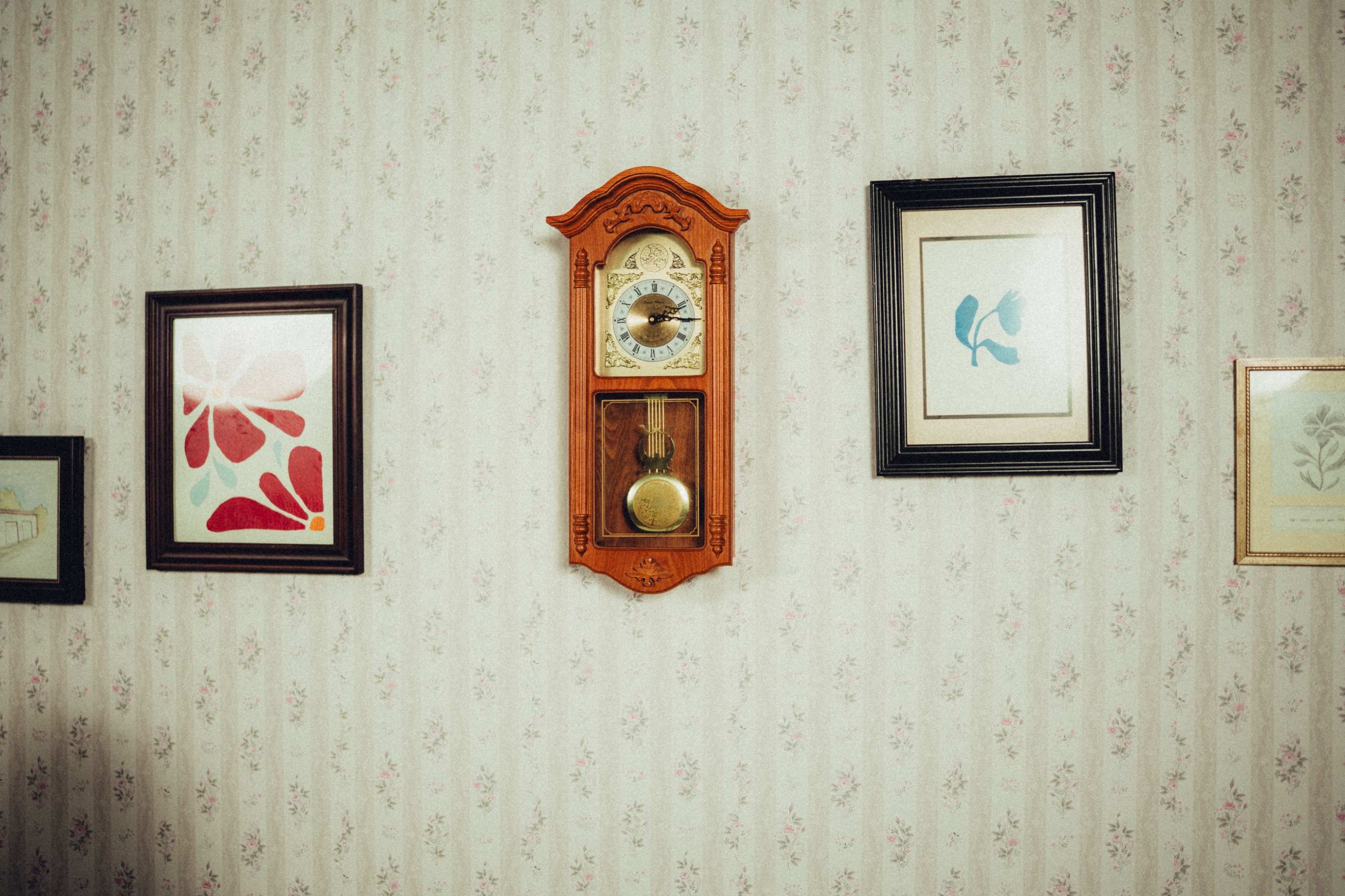 The image features a vintage wall clock centered between framed artworks on a patterned wallpaper background.