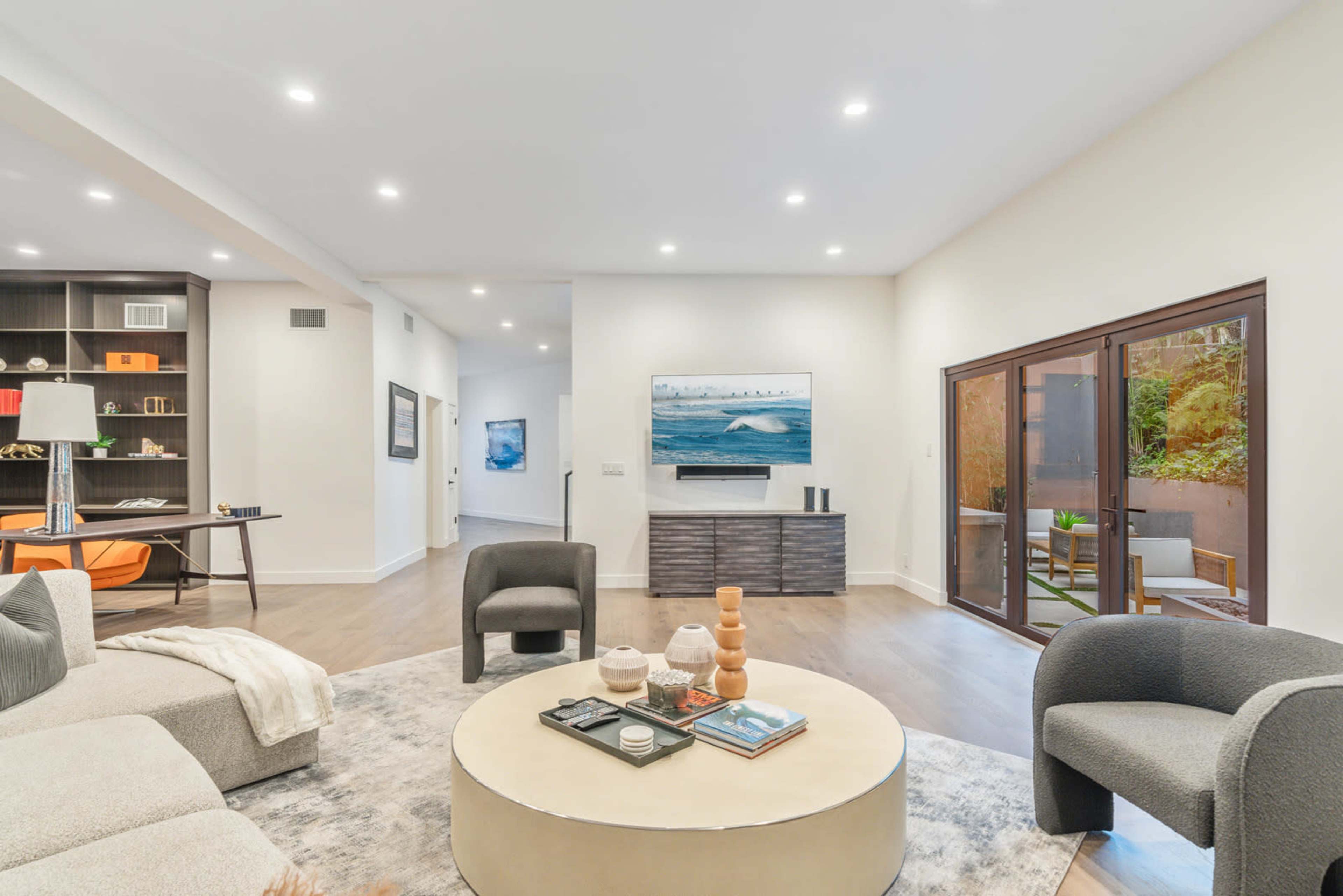 The image shows a modern, open-concept living room with a circular coffee table, a sectional sofa, and a television mounted on the wall, adjacent to large sliding glass doors leading to an outdoor area.