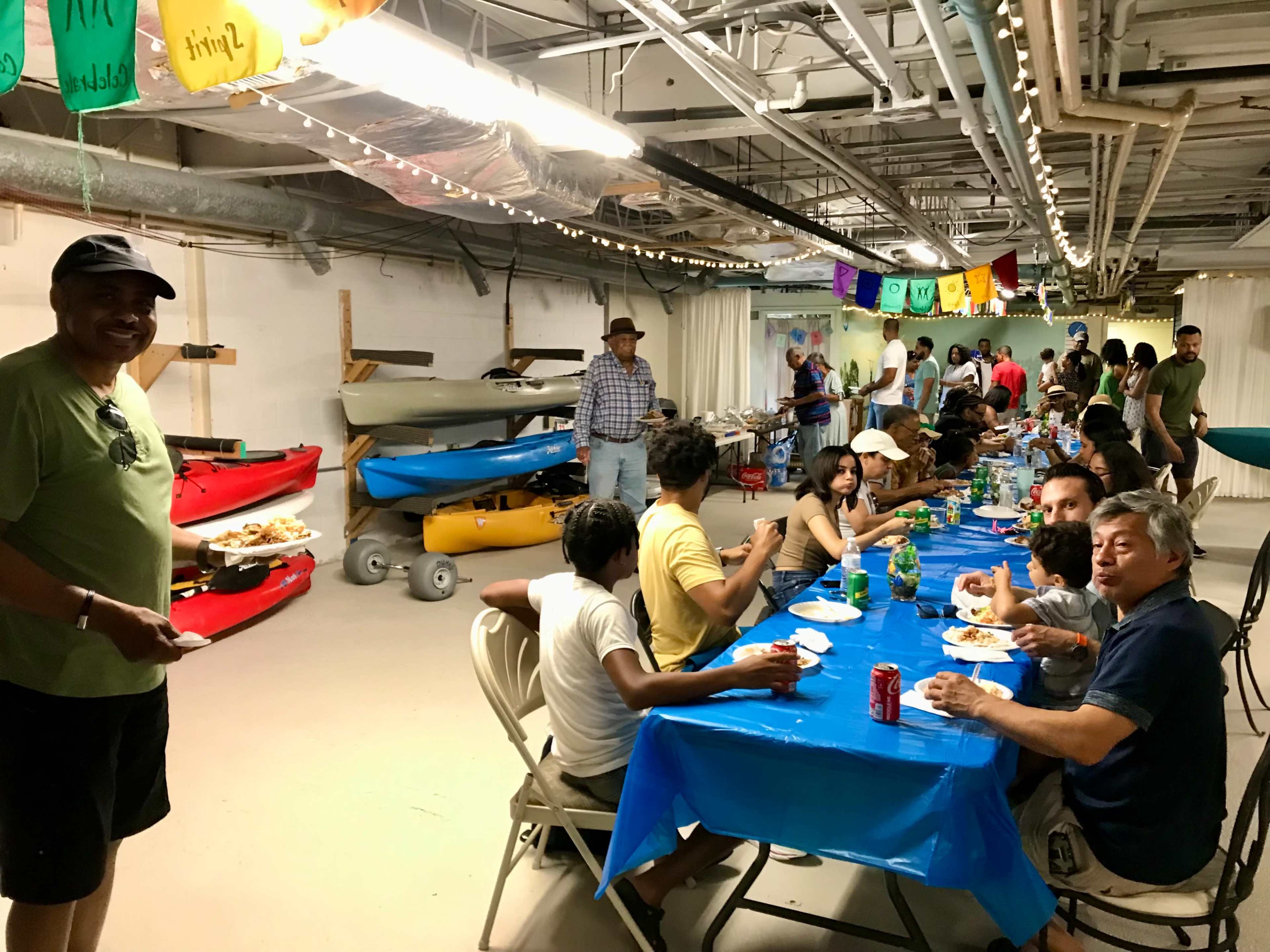 A diverse group of people gathers around a long table covered with blue tablecloths, eating and socializing in a room filled with kayaks and festive decorations.