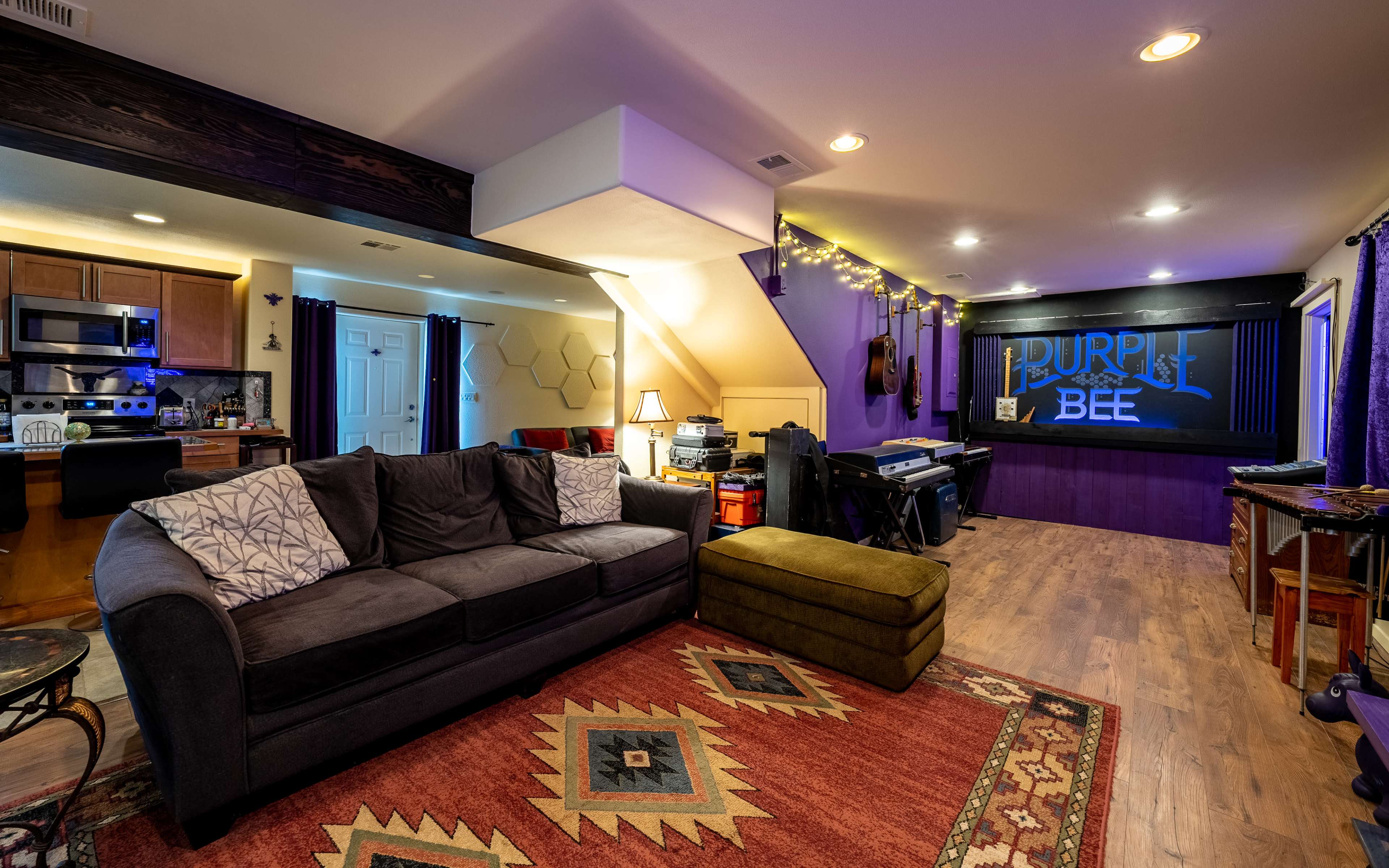 The image shows a cozy living space featuring a dark gray couch, a green ottoman, and a wall with "PURPLE BEE" signage, alongside a small kitchen area and musical instruments.