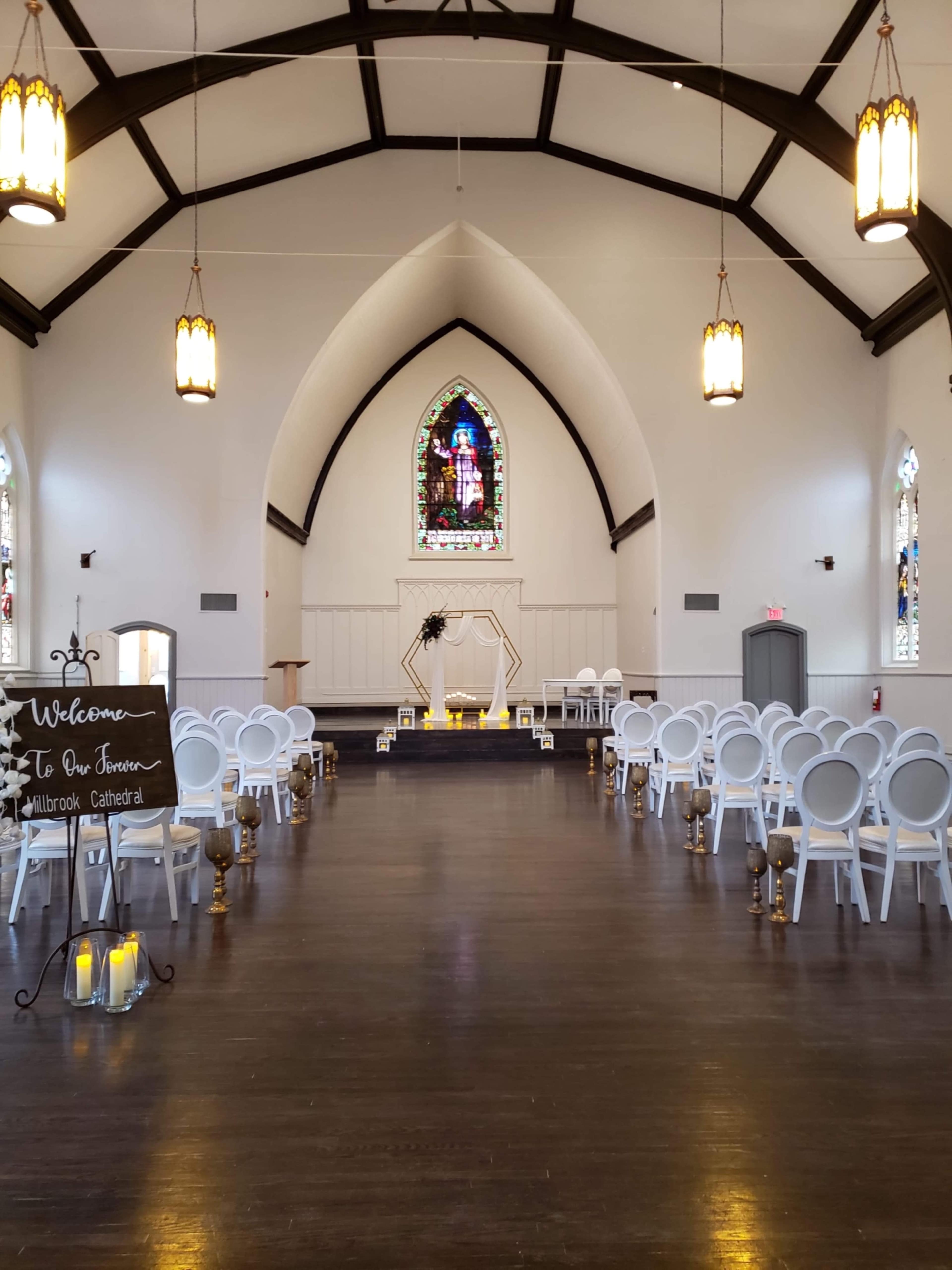 The interior of a church features rows of white chairs facing a decorative altar and a stained glass window at the far end.