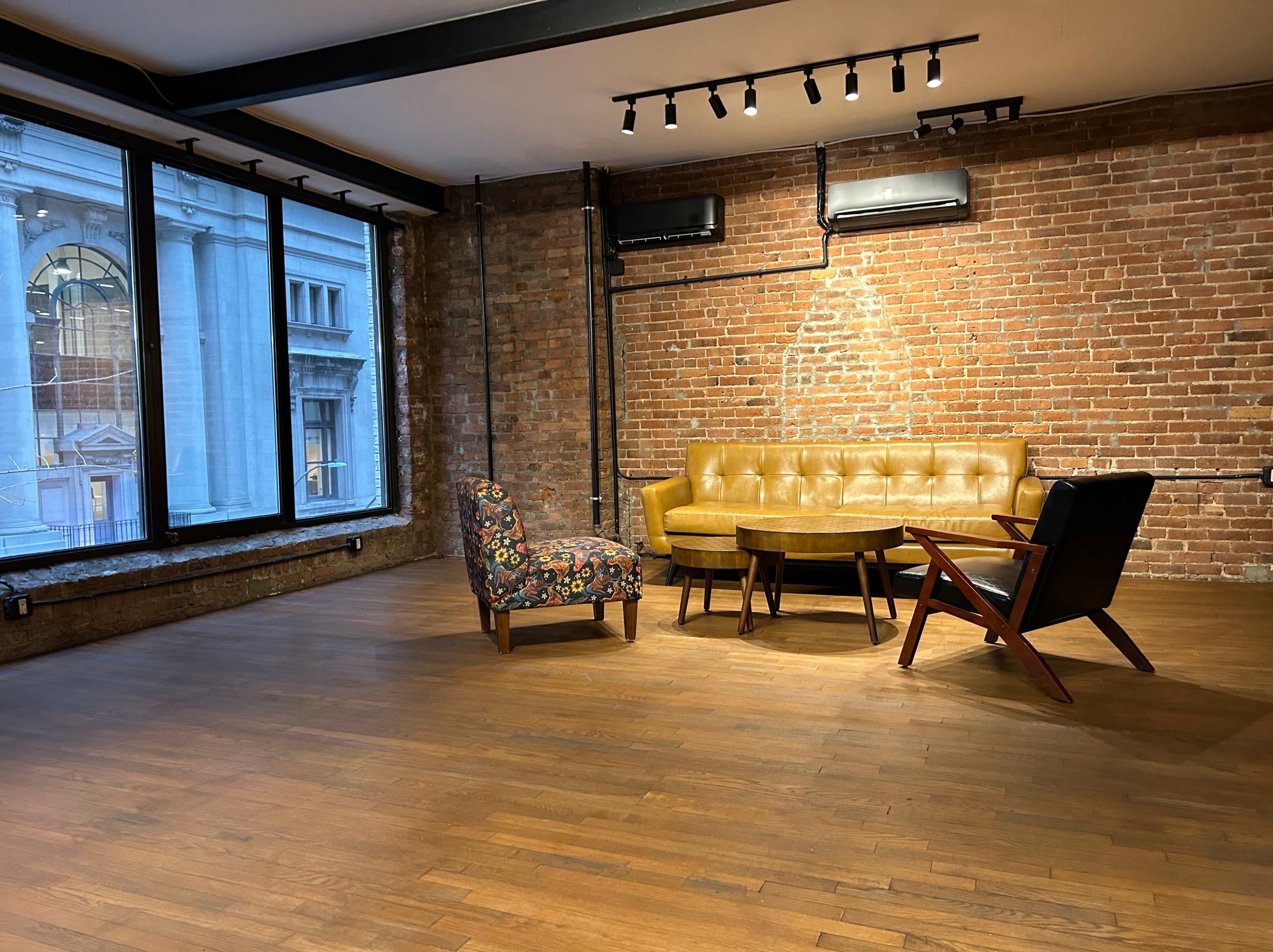 The image shows a modern interior space with a mix of seating options, including a yellow leather sofa, a patterned armchair, and a round table, set against a backdrop of exposed brick walls and large windows.