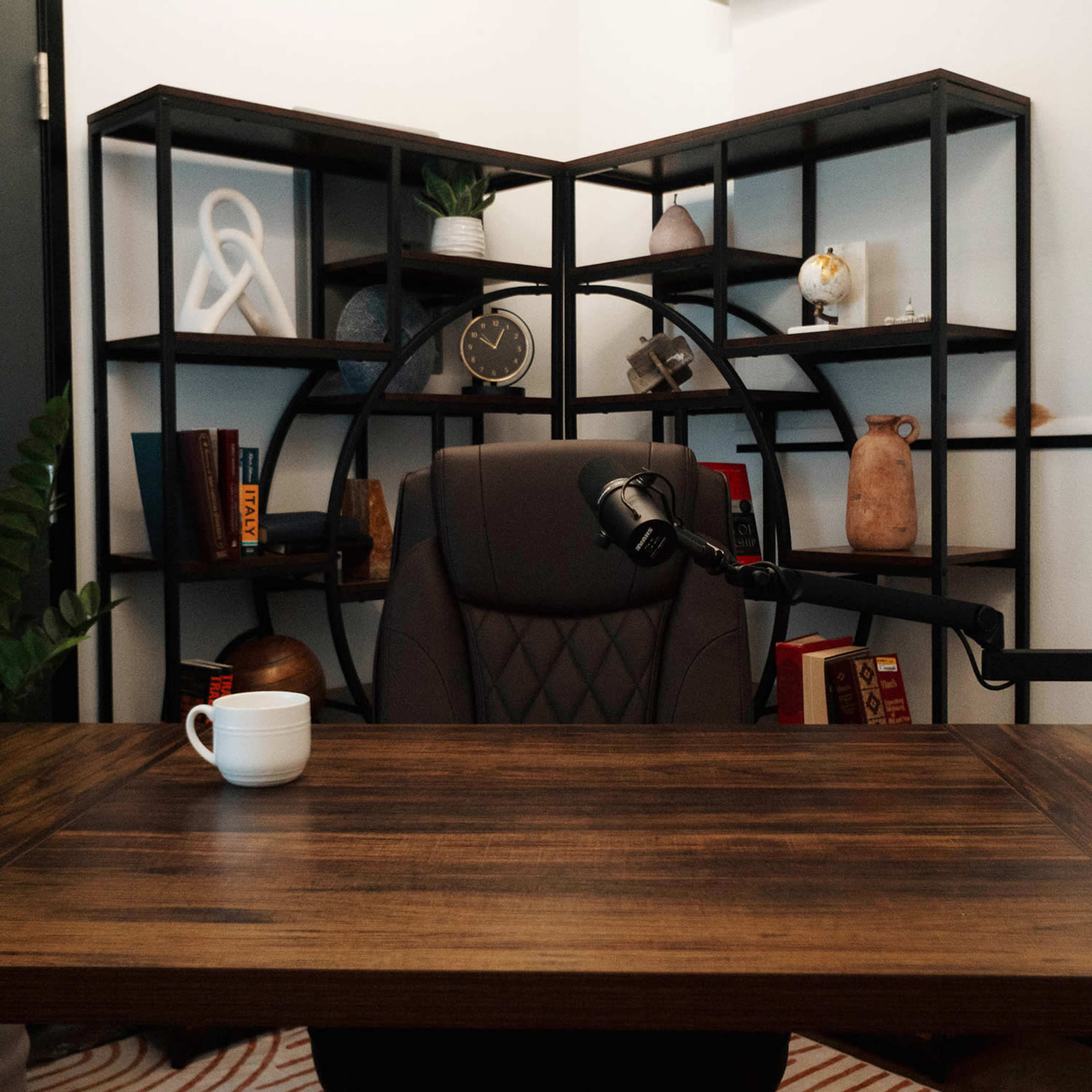 A modern office space features a wooden desk with a microphone, an empty chair, and a geometric bookshelf filled with various decorative items.