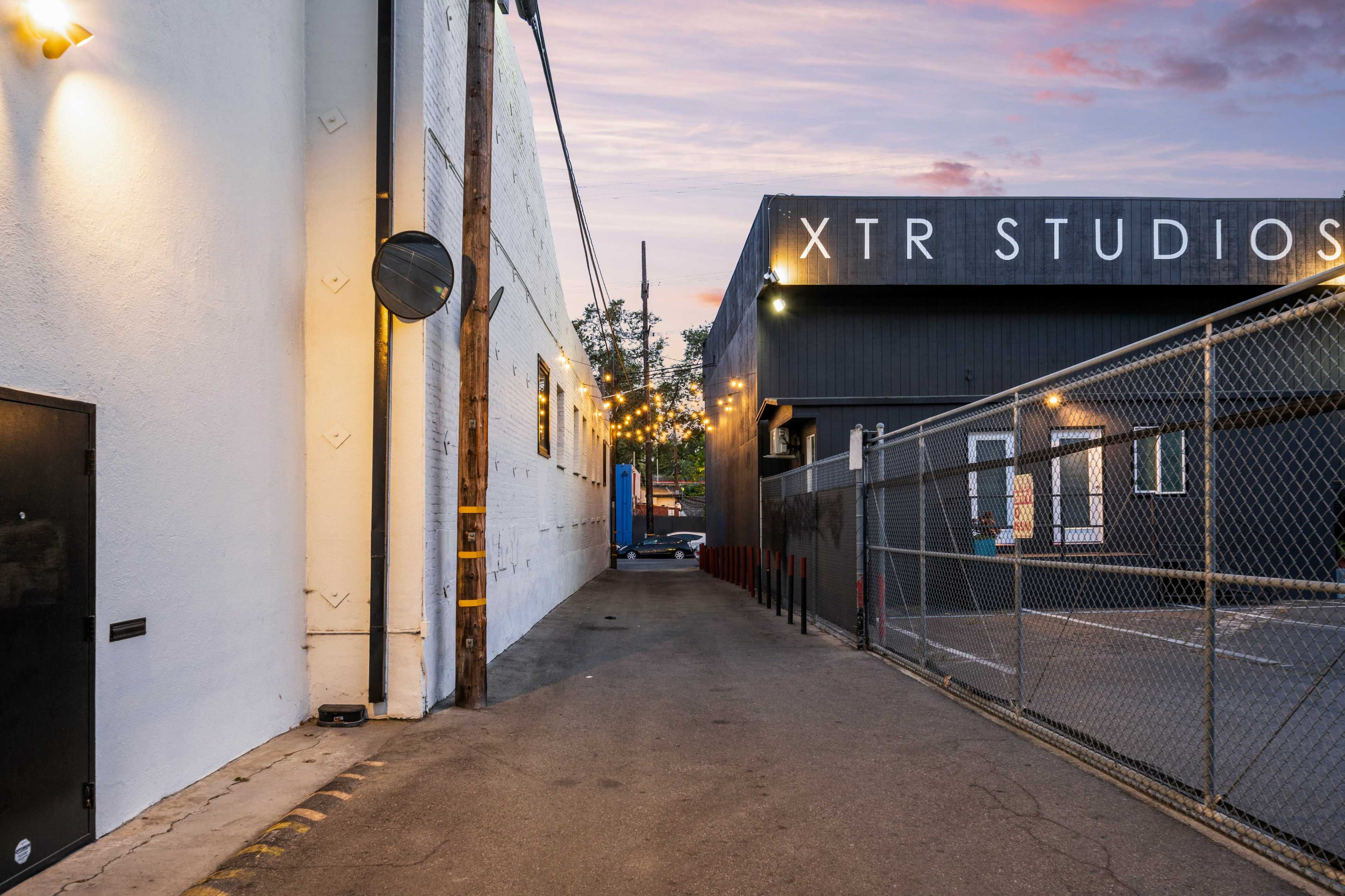 A narrow alleyway between two buildings, with a fence and string lights overhead, leading to a black structure labeled "XTR STUDIOS."