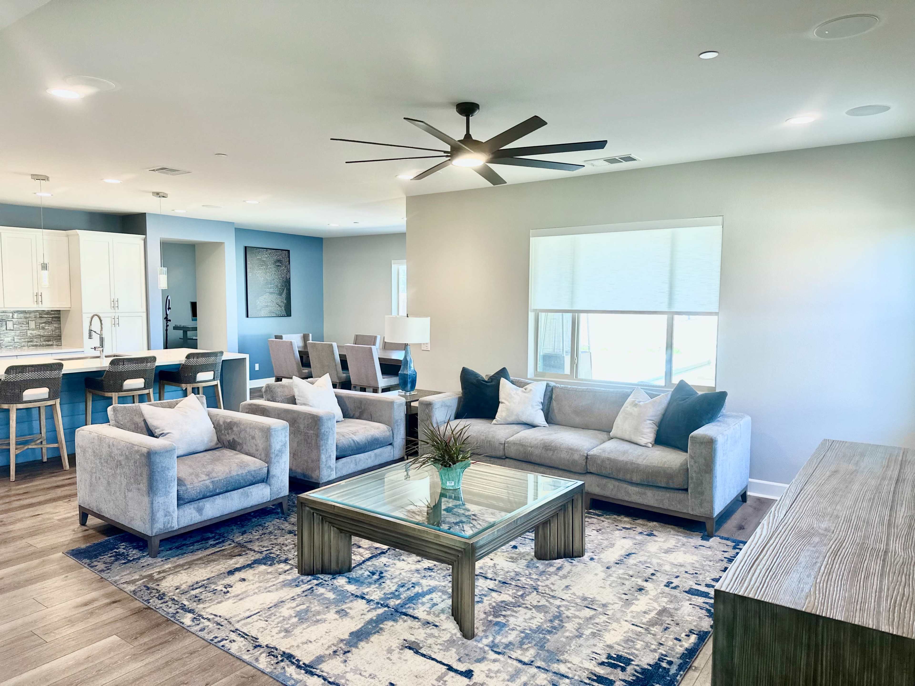 The image shows a modern living room with a gray sofa, two armchairs, a glass coffee table, and a large window, complemented by a kitchen area in the background.