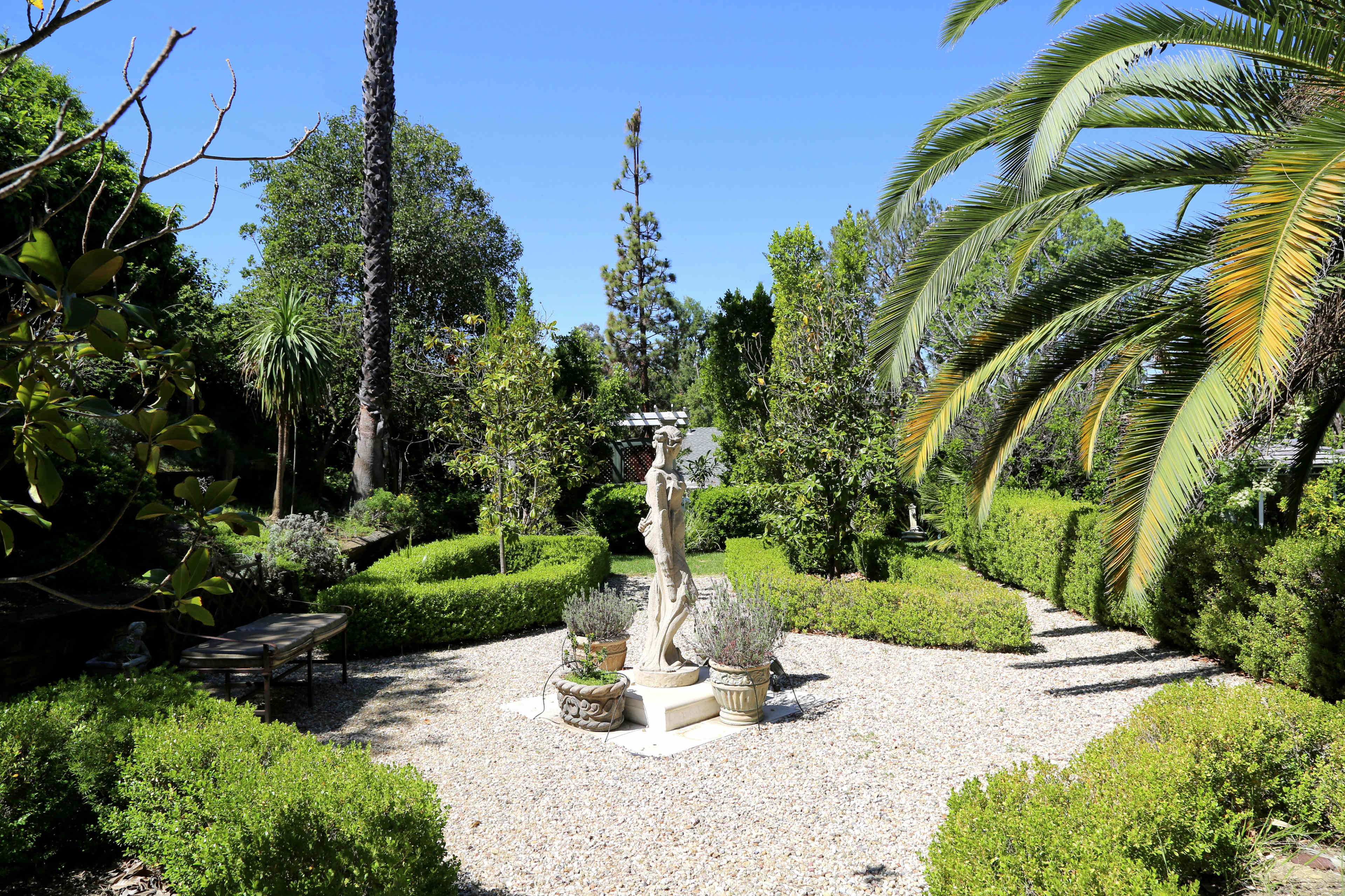 A landscaped garden featuring a central statue surrounded by neatly trimmed hedges and palm trees.